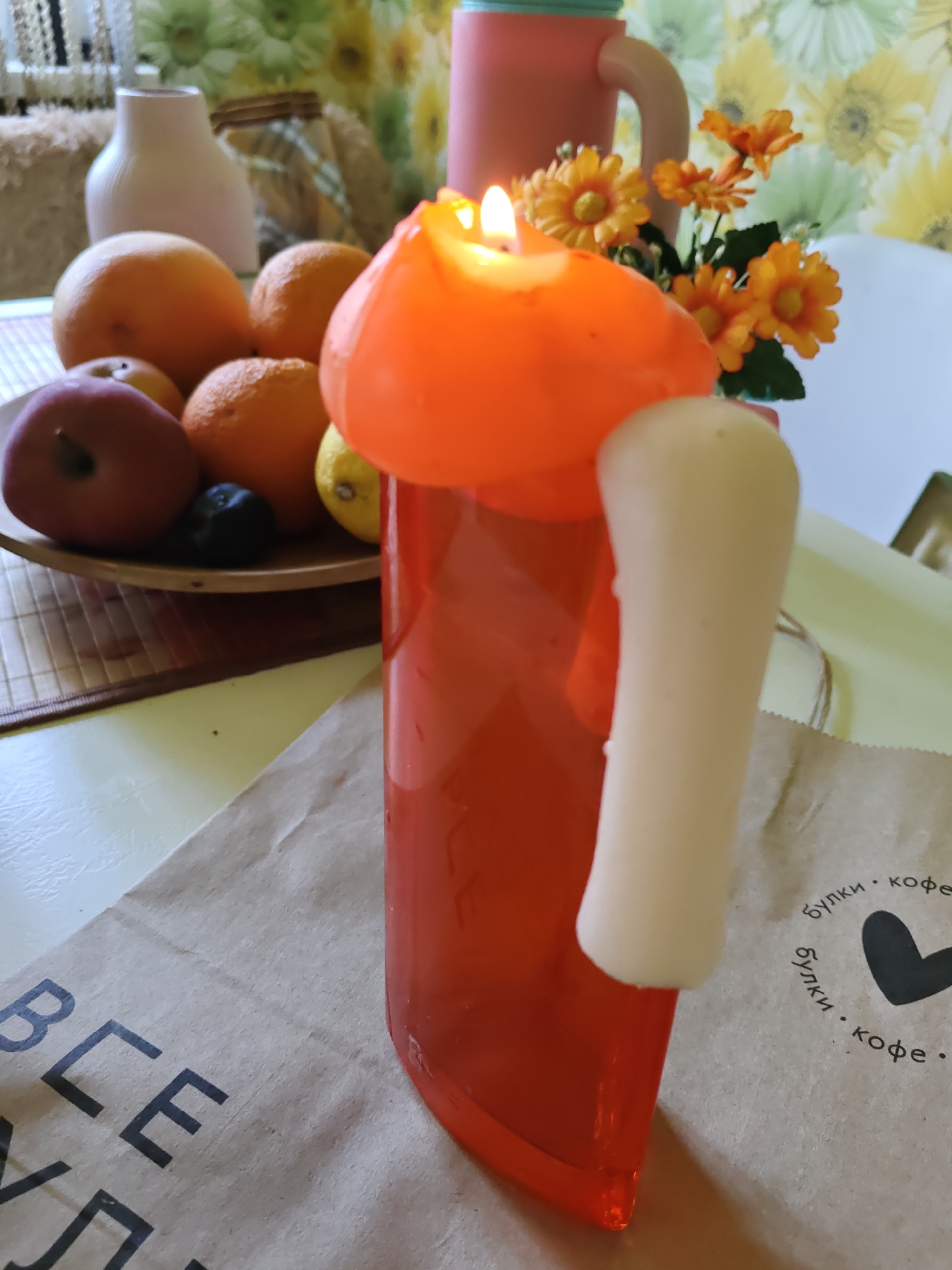 The image depicts a still life arrangement on a table, featuring a burning orange candle shaped like a persimmon, with a white cylindrical wax element attached to it. The candle sits in front of a glass vase filled with artificial orange flowers and greenery. To the left, a bowl contains various fruits, including oranges, a red apple, and what appear to be dark berries. Behind the fruit bowl, a white ceramic vase is visible, along with a patterned cloth. In the background, a pink mug with a beige handle stands out against a floral wallpaper. The foreground is occupied by a brown paper bag with black text and a logo consisting of a heart shape. The scene is set indoors, likely on a table, with natural light illuminating the objects. The provided crops offer closer views of specific elements within the arrangement. There is no information available to ascertain the location in the city of Rostov-on-Don, Russia, or any specific context beyond what is visually present in the image.