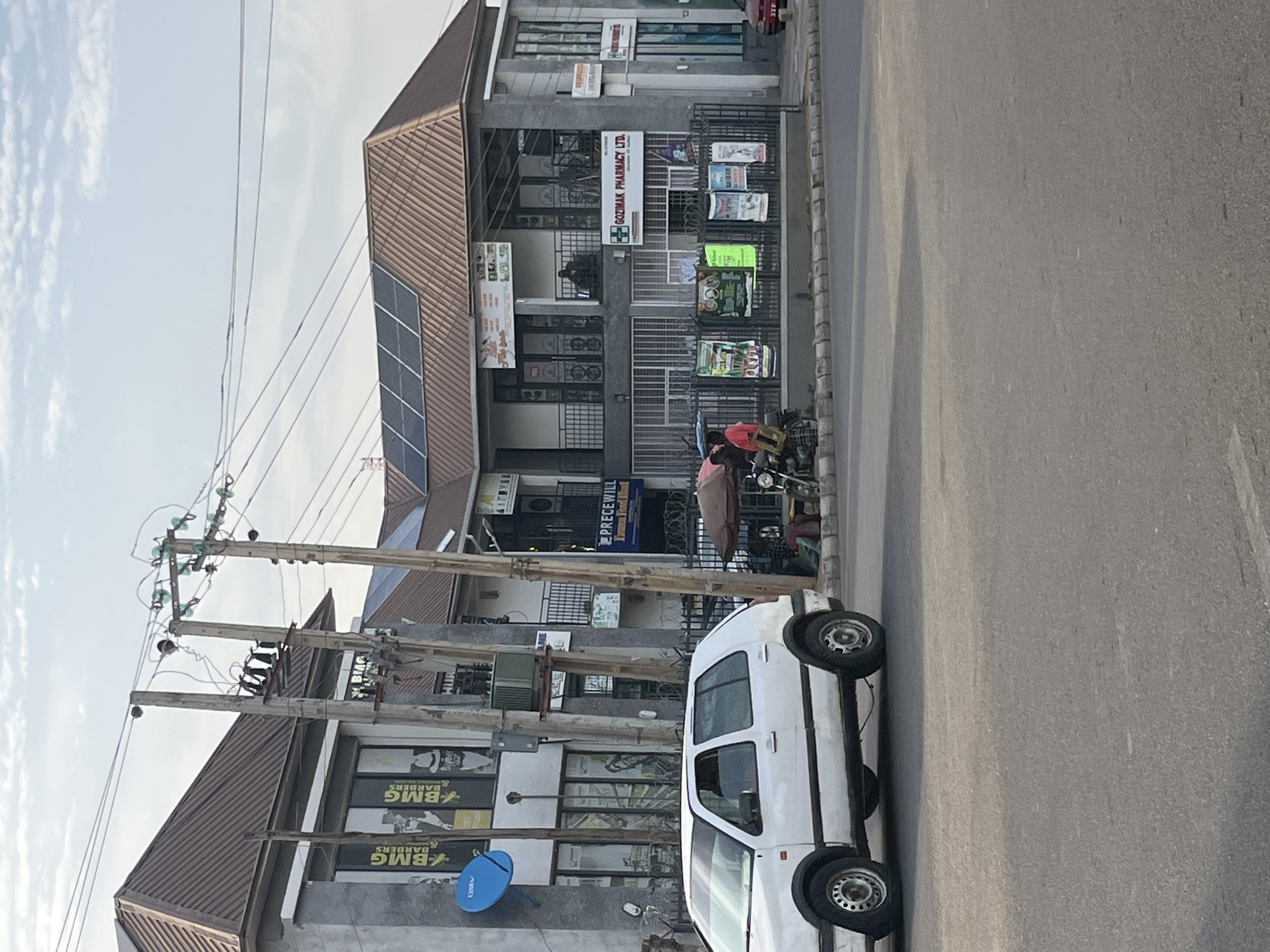 The media file displays an urban street scene, rotated 90 degrees clockwise. The scene features a row of two-story commercial buildings lining a paved road, under a bright, partly cloudy sky.
The buildings have brown, ribbed, sloped roofs, with solar panels visible on the roof of the leftmost building. Their facades are a light grey material, with ground-floor entrances and windows often protected by metal grilles. Several businesses are identified by their signage:
* "GOZEMAK PHARMACY LTD." is prominently displayed on the rightmost building. Below it, various smaller, colorful posters, some depicting food, are visible.
* Multiple signs reading "BMG BARBERS" are visible on the windows of the leftmost building.
* Another sign, partially obscured, appears to say "PRECEWILL."
A wide asphalt road occupies the foreground, with a white, compact van or utility vehicle parked on the side, near the leftmost building. A bright blue, circular satellite dish is affixed to the wall of this building. Along the street, multiple wooden utility poles, fitted with insulators and transformers, carry numerous power lines across the scene.
A few people are present: one person in light clothing stands near a motorcycle parked in front of the Gozemak Pharmacy, and another individual is visible sitting inside one of the shops.
The clear sky and bright illumination suggest the time of day is mid-morning to late afternoon. The scene depicts a typical commercial area with shops and parked vehicles.