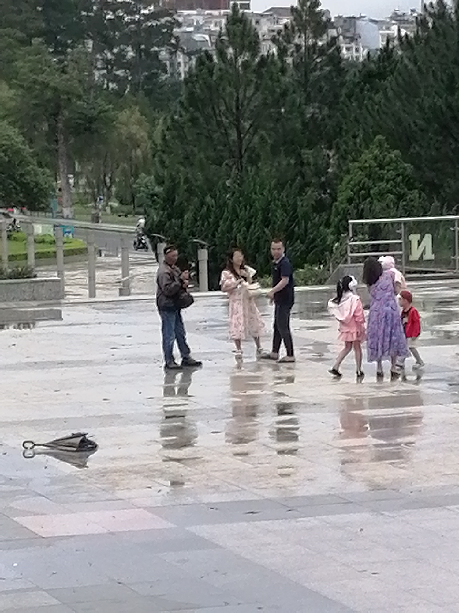 In Da Lat, Vietnam, on a wet, tiled plaza, a group of people stand around. A man in a dark jacket and blue jeans stands on the left, holding a phone. To his right, a woman in a light pink floral dress looks at the man. Next to her, another man in a dark blue shirt and dark pants stands facing the camera. Further right, a young girl in a pink jacket walks away from the camera, and a woman in a purple floral dress stands with a child in a red jacket to her right. A black umbrella lies on the wet ground in the foreground. The environment suggests an overcast day, possibly after rain.