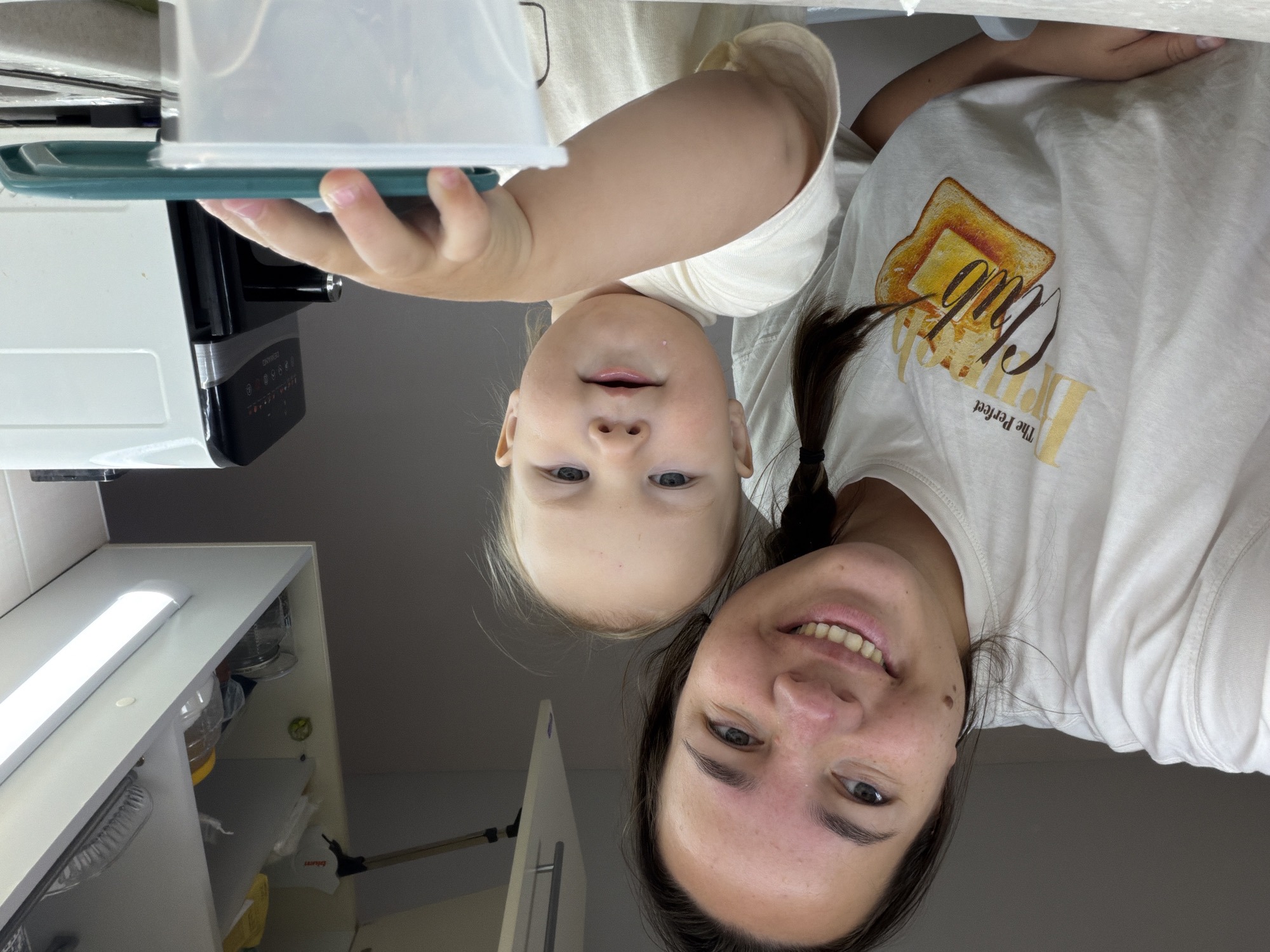 This image depicts a woman and a young child in what appears to be a kitchen environment. The woman is on the left, smiling and looking towards the camera. She has dark hair in a braid and is wearing a white t-shirt with a graphic of toast. The child is on the right, also looking towards the camera with a slight smile. The child is wearing a light-colored t-shirt and is reaching towards a green object on a white container. In the background, there are kitchen cabinets with some items on shelves, including jars and what looks like a pie dish. A white appliance, possibly an air fryer, is visible on the right side of the frame. The lighting suggests an indoor setting, possibly during the day given the bright overhead light. The scene appears to be domestic. There are no discernible landmarks or features that would indicate a specific location within Chaikovsky, Russia.