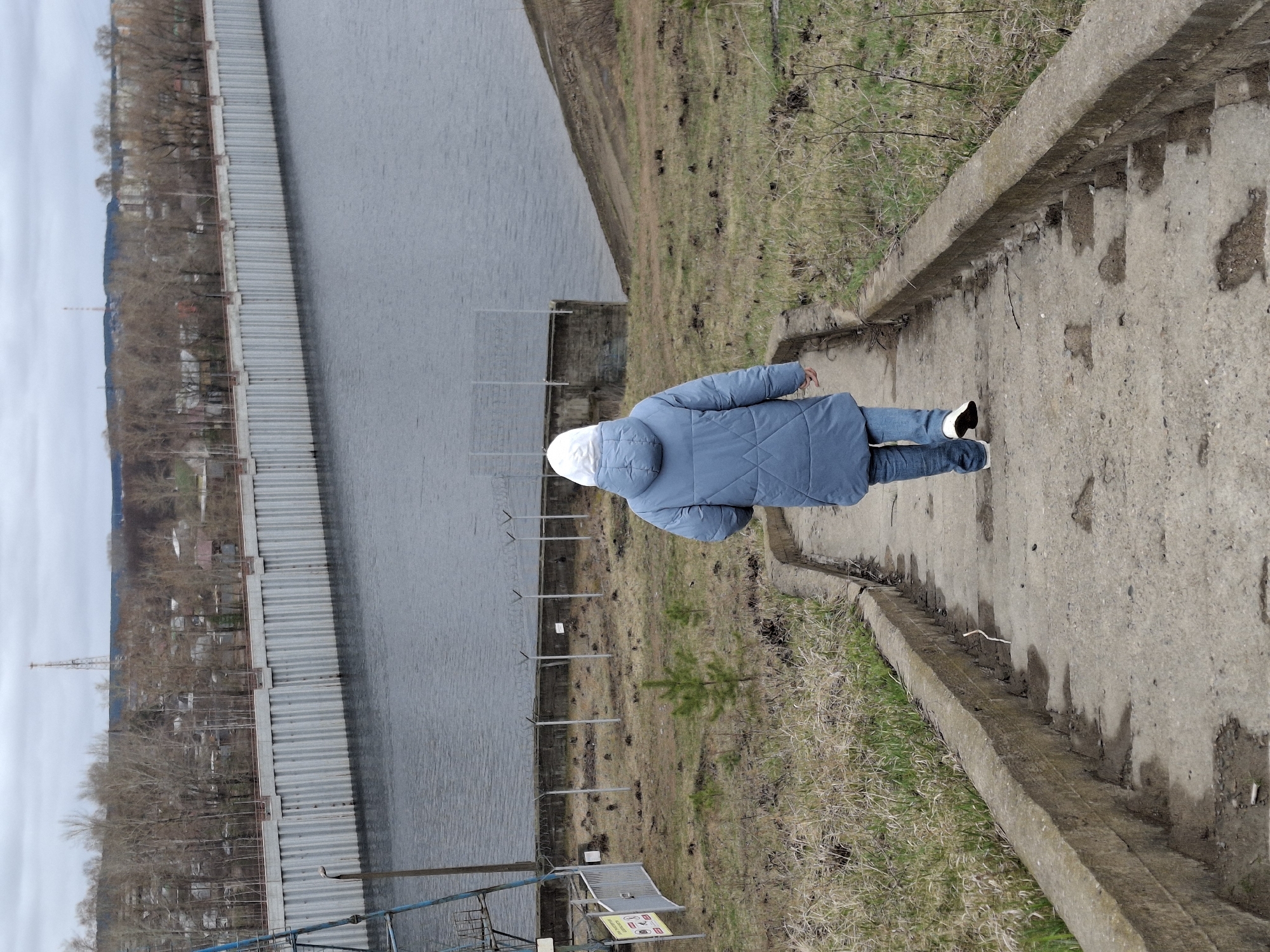 The image, taken in Chaikovsky, Russia, depicts an outdoor scene beside a large body of water, likely a river or reservoir, under an overcast sky. A person, viewed from behind, is walking down a concrete path or a series of rough steps. They are dressed in a light blue puffer jacket with a white hood, blue jeans, and black and white sneakers.
The concrete path features an elevated edge and descends towards the left. The ground immediately adjacent to the path consists of dry, patchy grass and dirt. To the left, a more overgrown grassy slope leads down to the water's edge.
The calm, grey body of water occupies a significant portion of the left frame. Along its bank, a long, corrugated metal sheet pile wall or similar barrier stretches into the distance. Beyond this structure, on the far bank, numerous bare trees line the shore, suggesting late winter or early spring. Small buildings are visible among the trees, and a tall communication tower stands prominently in the distant landscape against the light grey sky.
Near the bottom left, partially obscured by a metal fence, is a small white sign with red and black symbols and text. The visible text clearly states: "Опасно! Приближаться к воде запрещено!" (Dangerous! Approaching the water is forbidden!). Other symbols on the sign include prohibitions against swimming, diving, and littering. Several metal posts are driven into the ground near the water, possibly forming part of a safety barrier. A small evergreen sapling grows in the grassy area between the path and the water. The scene conveys a somewhat stark, possibly functional or semi-industrial riverside environment with explicit safety warnings.