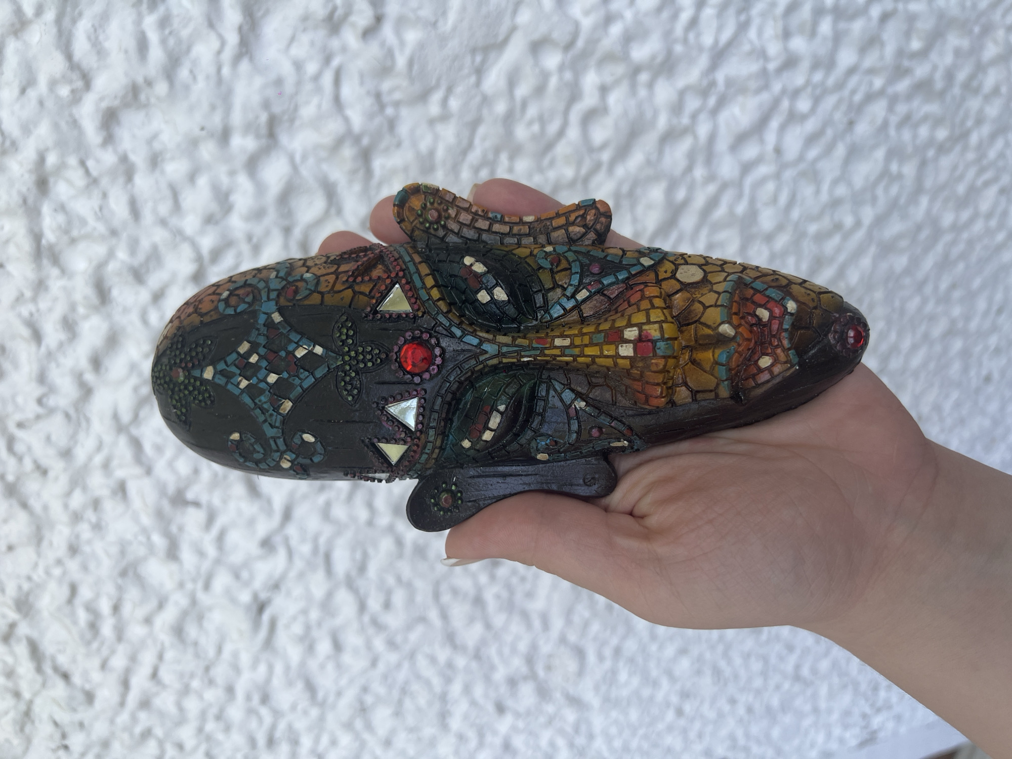 A person's hand is holding a decorative mask against a white textured wall. The mask is elongated and somewhat oval-shaped, with a dark base color and intricate mosaic-like patterns in shades of blue, red, yellow, and brown. It has embedded gemstones or reflective pieces in red and silver. The location is not discernible from the image.