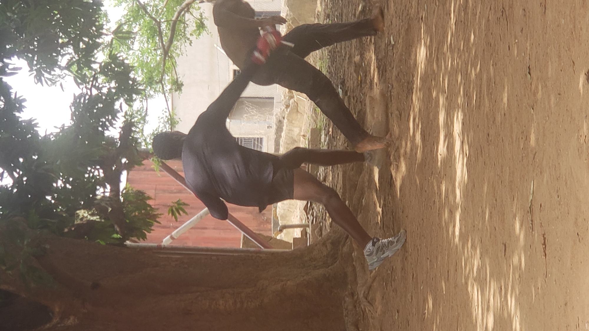 In an outdoor setting in Narayi, Nigeria, two males appear to be engaged in a physical altercation. One male is wearing dark shorts, a dark shirt, and one shoe. The other male is wearing dark pants and is shirtless. Both are dark-skinned. The setting includes a dirt ground, trees, a building with windows, and what appears to be a red barrier with white piping. The scene is well-lit and taken during the daytime.