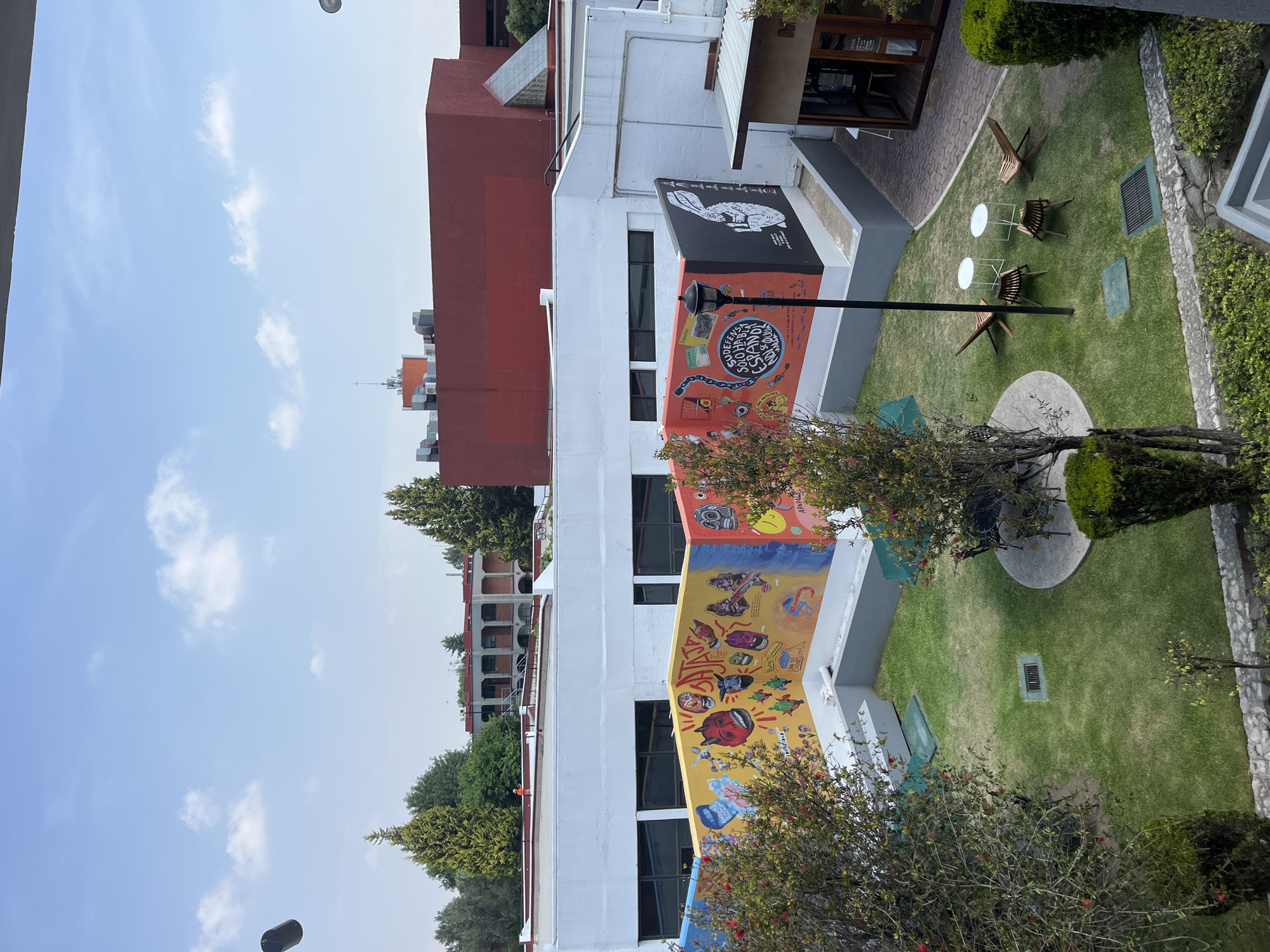 The image captures a sunlit outdoor courtyard or garden area, likely situated in San Andrés Cholula, Mexico, as indicated by the context. A clear blue sky with sparse, fluffy white clouds dominates the upper half of the scene, suggesting bright daylight, possibly midday or early afternoon.
The main subject is a multi-story white building on the right, distinguished by a long, colorful mural spanning much of its exterior facade. The uppermost section of the mural features a prominent black and white illustration of a rabbit. Below this, various vibrant panels depict diverse artistic and cultural motifs. Text visible on the upper mural includes "INDEFENSA SOY CUANDO ESTOY EN TUS MANOS" and the word "Español," along with "CUADRADO DE LIBROS." Further down the mural, phrases such as "AQUI SOY LIBRE" and "LANZADA...CHOLULA" can be partially read amidst other intricate designs.
Adjacent to the building is a manicured green lawn, furnished with several modern wooden lounge chairs and small, white circular tables, suggesting a welcoming and casual outdoor seating area. A black lamppost stands near the building and the lawn. On the grass, a circular concrete patio feature and a rectangular metal grate are visible.
In the background, a reddish-brown building with a flat roof and industrial equipment is visible behind the white structure. Further to the left and in the distance, another building with multiple windows, possibly featuring arches or balconies, is partially obscured by lush green trees. Additional trees and bushes frame the foreground, contributing to the serene and verdant atmosphere of the space. No people are present, emphasizing the tranquil and inviting nature of the setting.
