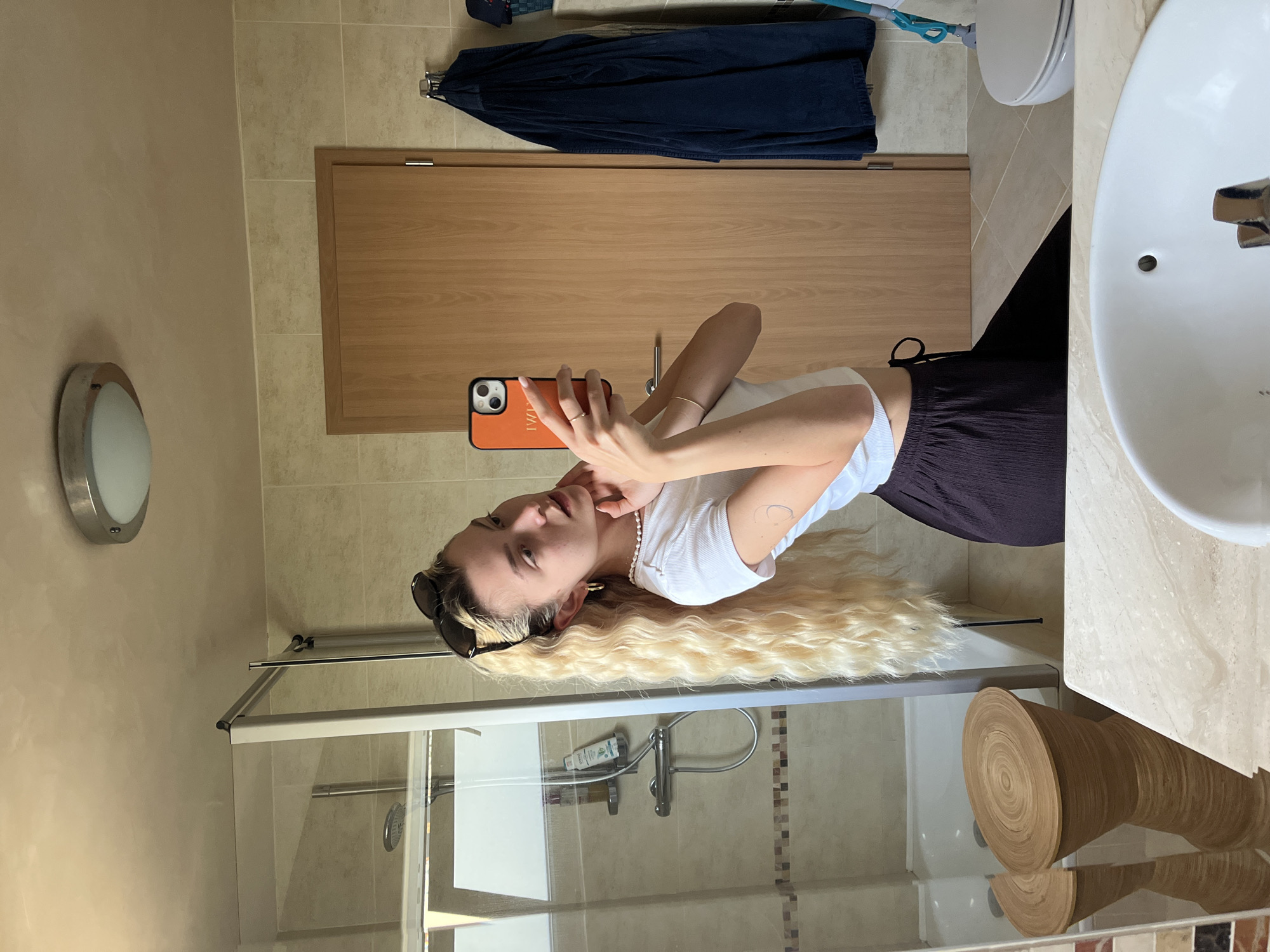 A young woman is captured in a mirror selfie inside a well-lit bathroom. She has long, wavy blonde hair with darker roots, and is wearing sunglasses pushed up on her head. She is dressed in a white ribbed crop top and dark, possibly pleated, trousers or skirt. Her left arm features a delicate bracelet and a small, light-colored tattoo. She wears a ring on her left ring finger and a pearl necklace around her neck. Her right hand holds an orange-cased smartphone, which displays the text "KIMI" on its back, capturing her reflection. She looks towards the phone with a neutral expression.
The bathroom features light-colored, possibly beige, tiled walls and floor. To the right, a white ceramic sink with a chrome faucet is visible, with the brand "SANINDUSA" faintly inscribed on it. Below the sink is a light-toned countertop. A wooden, cylindrical stool or side table with stacked circular layers is reflected in the lower right.
In the background, there's a shower enclosure with clear glass doors and chrome framing. Inside the shower, various shampoo bottles and a shower head can be seen. To the left of the shower, a circular wall-mounted light fixture is present. A light wooden door with a horizontal metal handle is visible behind the woman. Above the door, a dark blue towel or garment hangs on a hook, alongside a lighter blue object. The overall setting is clean and modern.