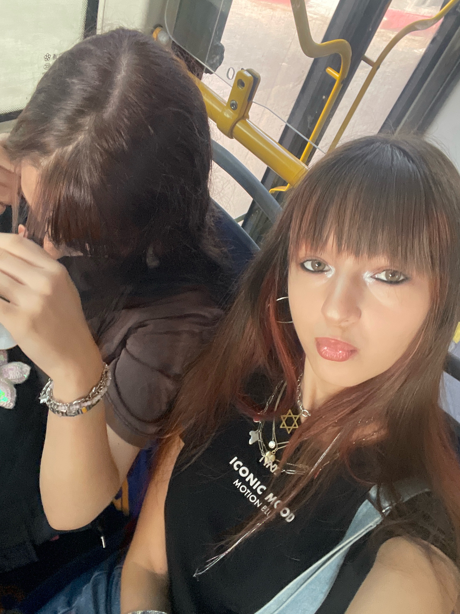The image shows two individuals seated inside a public bus. The person in the foreground, facing the camera, has long brown hair with bangs, brown eyes with dramatic eye makeup, and is wearing a black t-shirt with the words "ICONIC MOOD MOTION BLU" printed on it, along with several layered necklaces. The person in the background, partially obscured, also has dark hair. Both individuals are wearing jewelry on their wrists. The environment suggests an indoor setting with bus interior elements like yellow grab bars and seating visible. There is no specific indication of the city of Raanana, Israel, within the image itself.