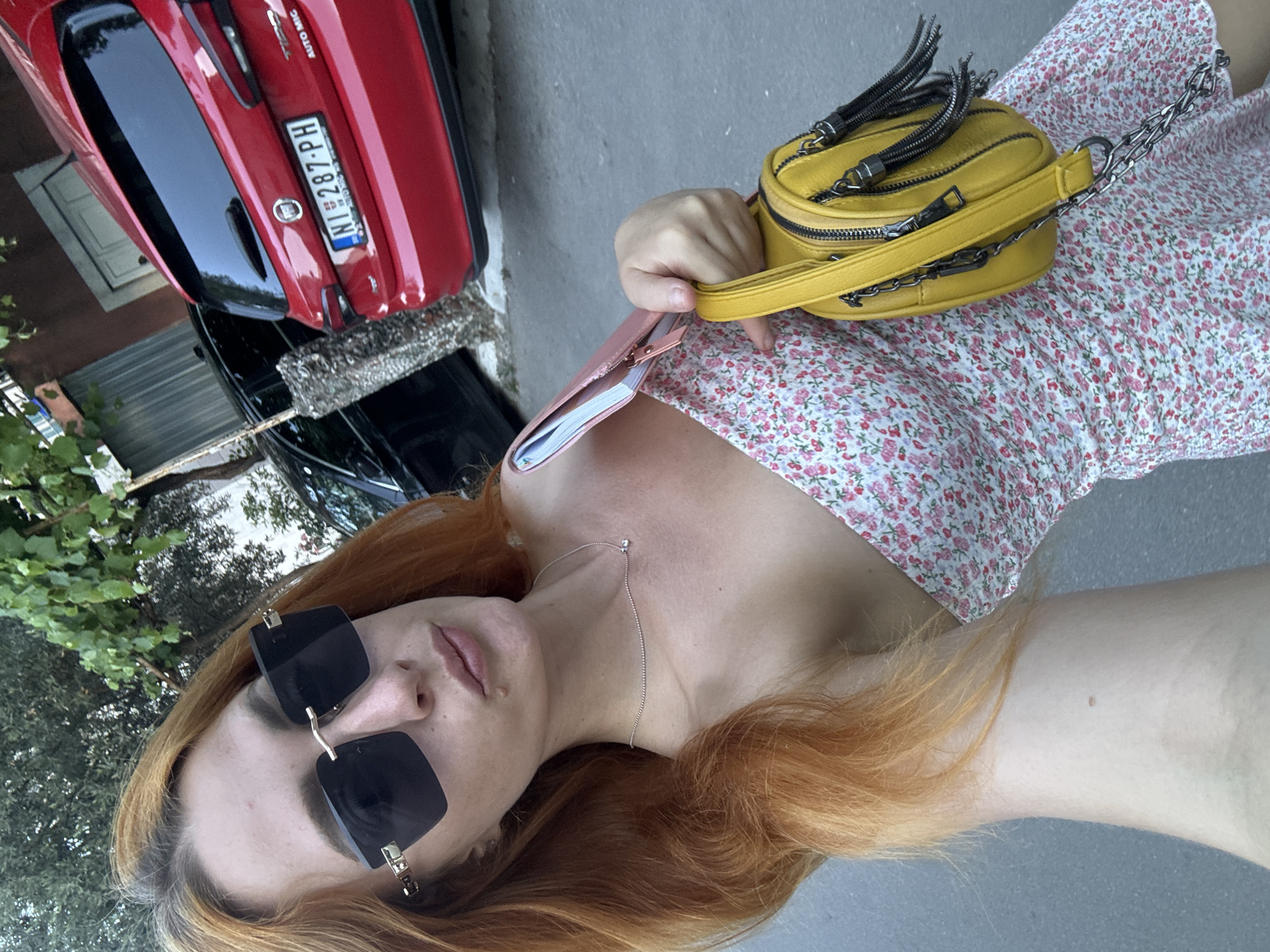 In Budva, Montenegro, a young woman with auburn hair and a floral print dress poses for a selfie. She is wearing large, dark sunglasses and a delicate silver necklace. A bright yellow purse with silver chain detailing hangs from her left shoulder. In the background, a red Fiat Tipo with a license plate that reads "NI 287-PH" is parked. A black car is visible next to the Fiat. The scene appears to be an outdoor urban setting with trees and a paved surface.