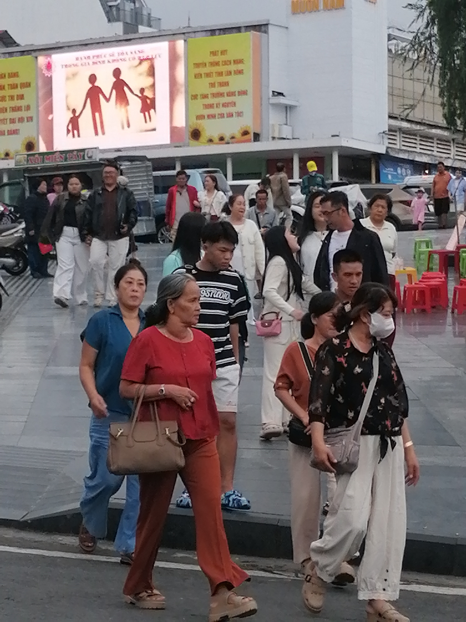 The image captures a street scene in Da Lat, Vietnam, likely in the evening or overcast conditions, given the lighting. In the background, a large building with a white facade is visible, featuring prominent signage with text and graphics, including an illuminated billboard displaying a silhouette of a family. To the left of this building, another billboard with Vietnamese text and sunflower imagery is partially visible. A group of people are walking across a paved area, which appears to be a pedestrian walkway or street.
In the foreground, several individuals are captured in mid-stride. A woman in a red top and orange pants, carrying a tan handbag, is prominently featured. Beside her, a woman in a blue shirt and jeans is walking. Further ahead, a man in a black and white striped shirt and shorts is walking. To the right, a woman wearing a white mask and a dark patterned top is walking. Other individuals are visible in the mid-ground and background, including people in casual attire, some carrying bags. Red and green plastic stools are arranged to the right of the frame, suggesting a nearby seating area or vendor. The ground surface is paved with grey tiles, and a slightly elevated dark grey curb is present. Vehicles, including motorcycles and cars, are partially visible in the background, indicating a public urban setting.