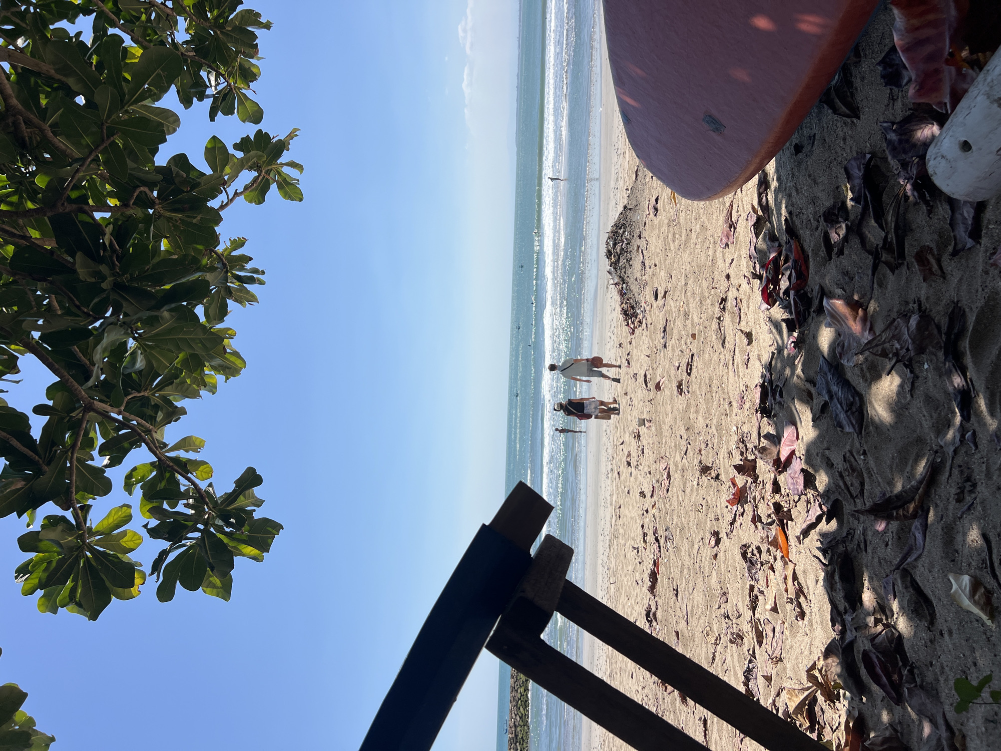 A bright, sunny day at Kuta Beach, Indonesia, is depicted in this eye-level shot. The foreground is dominated by the sandy beach scattered with dry leaves, and the edge of a red surfboard is visible on the right. A dark, wooden structure, likely a beach chair or shelter support, juts in from the left foreground, casting a shadow. In the middle ground, two figures, a male and a female, walk towards the ocean. The woman wears a white top and patterned shorts, carrying a brown bag, while the man is in a light-colored t-shirt and shorts. Further out, several other individuals can be seen wading in the gentle ocean waves, with more people visible further out in the water, some appearing to be surfers or swimmers. The ocean is calm with small waves breaking, and the sky is a clear, vibrant blue with a few wispy clouds near the horizon. A breakwater made of rocks is visible on the left side of the image, stretching out into the sea. The overall scene is one of a relaxed beach day.