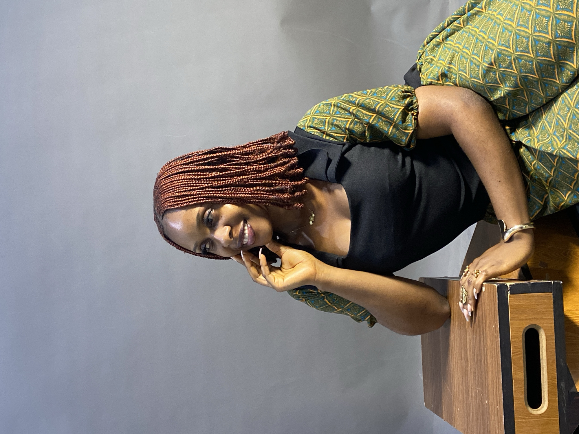 A medium shot shows a brown-skinned woman with reddish-brown braided hair, wearing a black top with green and gold patterned sleeves, leaning on a wooden box with her right hand and fingers touching her face. She is smiling and looking towards the viewer. She is wearing a necklace, rings, and a bracelet. The background is a plain gray wall. There is no information to indicate the location is in Jos, Nigeria.