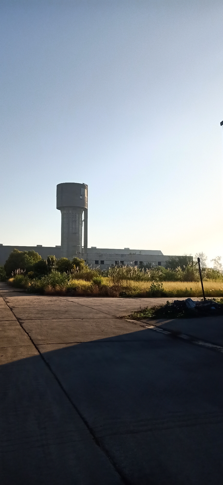 The image captures a desolate industrial landscape under a clear, pale blue sky. In the background, a large, imposing concrete water tower stands tall, its cylindrical upper section distinct from the conical base. Adjacent to the tower is a long, low-lying industrial building with numerous windows. The foreground is dominated by a paved area, possibly a road or courtyard, marked with concrete seams and casting a long shadow to the right, indicating the sun is low in the sky. Overgrown vegetation, including tall grasses and scattered bushes, flanks the building and extends towards the paved area, suggesting a lack of recent maintenance. The overall impression is one of abandonment and neglect. The location is identified as Keudekruenggeukueh, Indonesia.