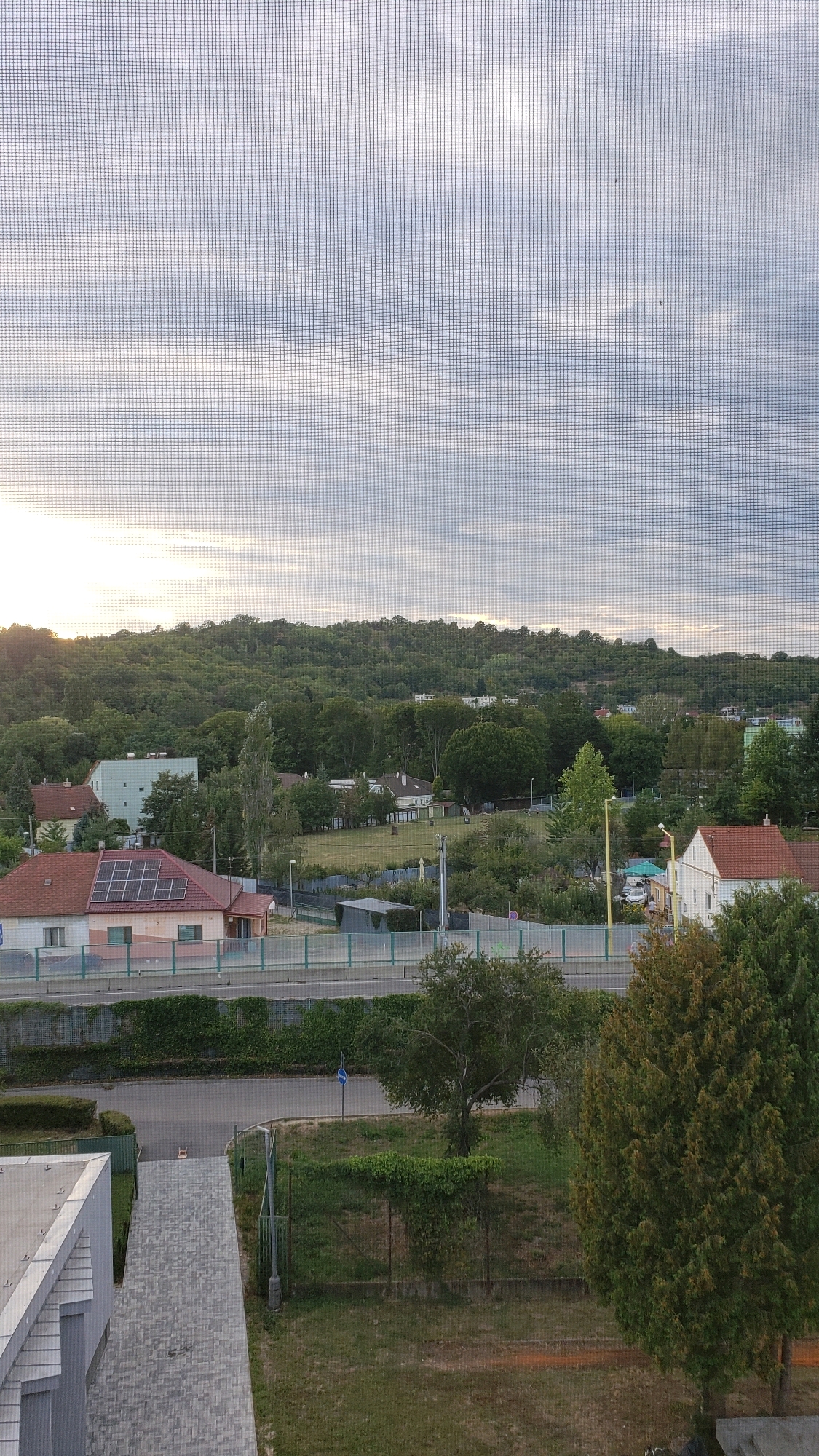 The image depicts a residential area in Prešov, Slovakia, viewed through a window screen. In the foreground, a gray modern building with a slanted roof is visible on the left. A paved pathway leads away from it. In the middle ground, a road runs horizontally, bordered by a fence with green ivy and a low hedge. Beyond the road, several houses are nestled among trees, with one house in the foreground on the left featuring solar panels on its red-tiled roof. The background is dominated by a lush, green, tree-covered hill. The sky is cloudy with streaks of sunlight, suggesting either early morning or late afternoon. There are no people visible in the scene. The overall impression is one of a quiet, suburban or semi-rural landscape.