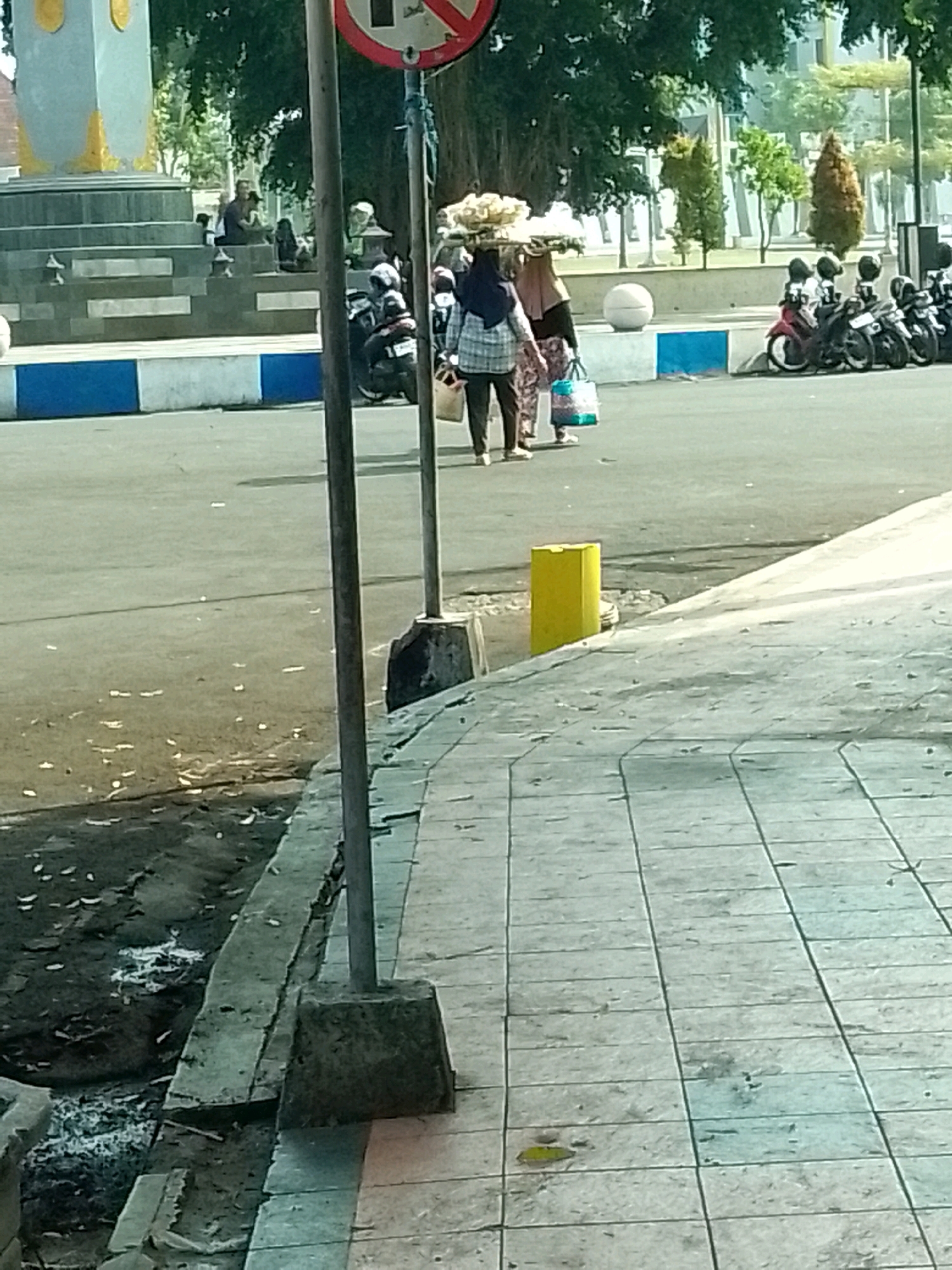 The image depicts an outdoor scene in Jember, Indonesia, likely near a public monument as suggested by the stepped structure and the presence of a traffic sign. In the foreground, a tiled sidewalk is visible, with a drainage ditch running alongside it. A metal pole with a concrete base stands on the sidewalk.
Further back, a street with parked motorcycles can be seen. Two women are walking on the street; one is carrying a large basket of food items on her head and a bag in her hand. The other woman is carrying a tote bag. The street is lined with blue and white barriers. In the background, trees and buildings are visible, indicating an urban or semi-urban environment. The overall lighting suggests it is daytime.