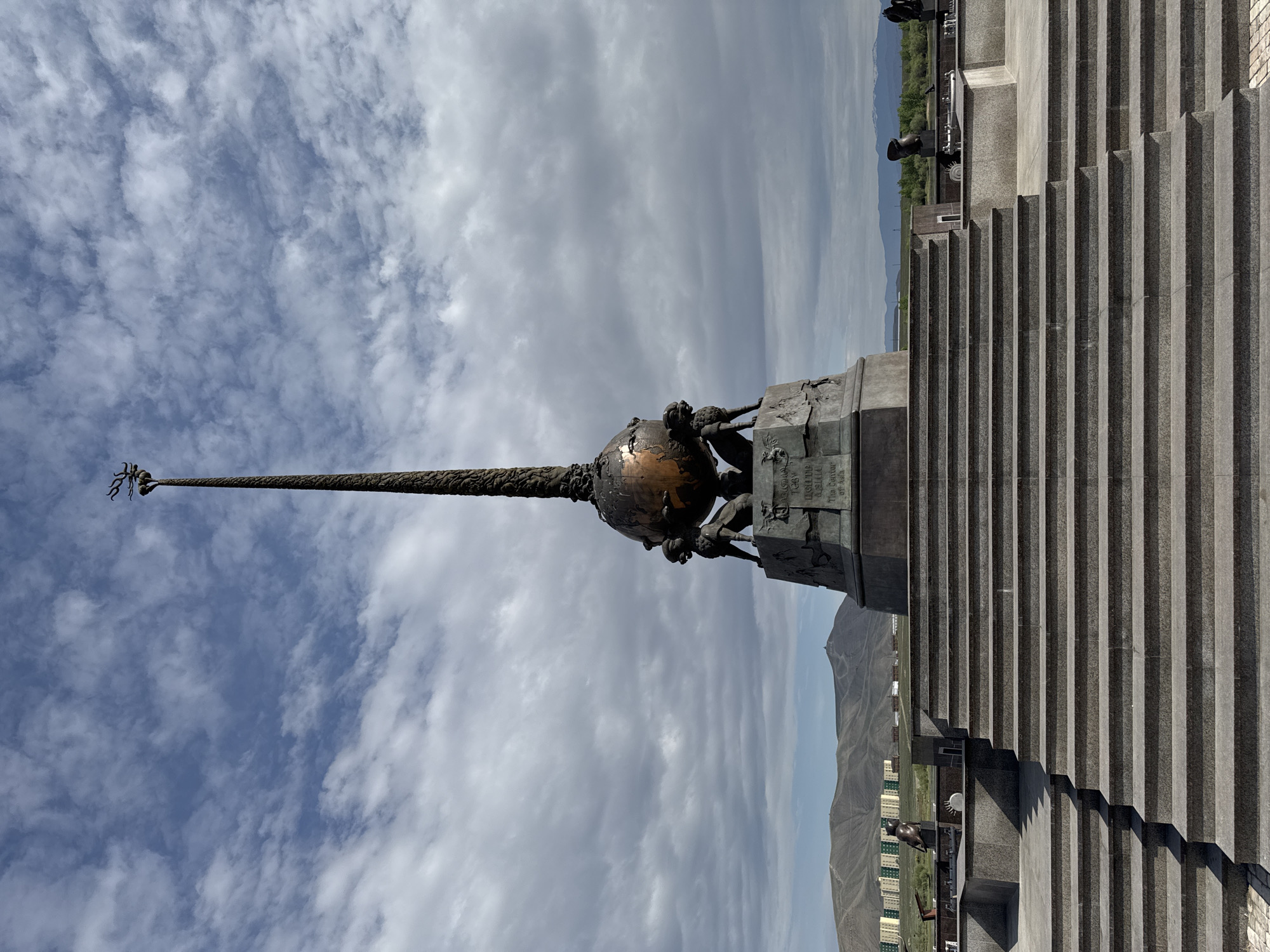 The image depicts the Center of Asia monument in Kyzyl, Russia. The monument consists of a bronze globe held by three supporting figures atop a stone pedestal. A tall obelisk rises from the globe, terminating in a stylized crown. The background features a cloudy sky, the Yenisei River valley, and distant mountains. A large staircase leads up to the monument's base. Several cannons are also visible on the upper part of the stairway. Modern buildings can be seen nestled in the valley below.