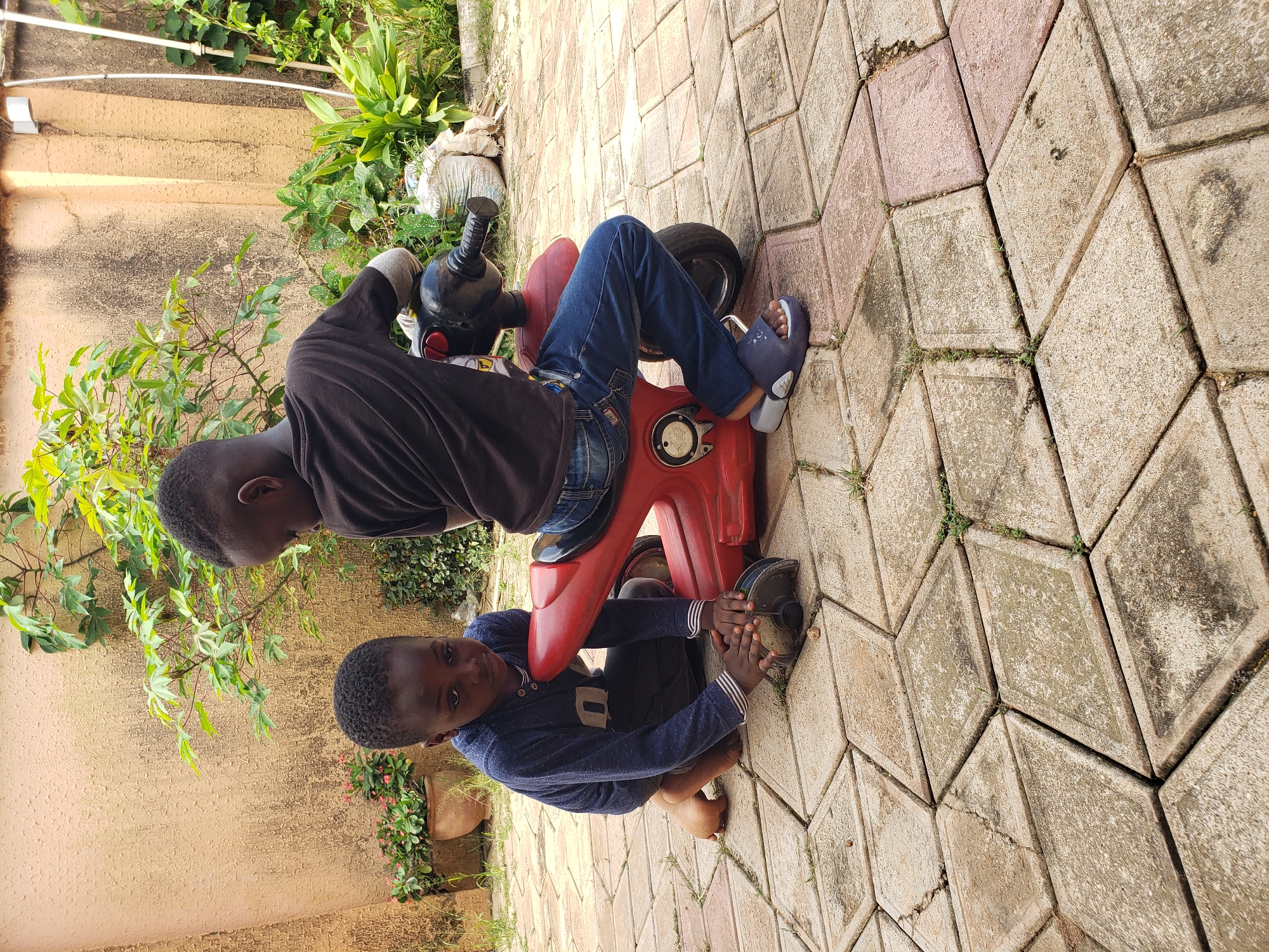 Two young boys are in an outdoor courtyard in Sabon Tasha, Nigeria. The boy on the left, wearing a navy blue long-sleeved shirt and dark pants, kneels on the diamond-patterned interlocking brick ground, looking intently at the front wheel of a red toy motorcycle. The boy on the right, dressed in a black t-shirt and blue jeans, sits on the red toy motorcycle, looking down. The courtyard has a textured, beige-colored wall in the background, partially covered with green foliage from a bush. In the distance, more green plants and a white sack are visible. The lighting suggests it is daytime.