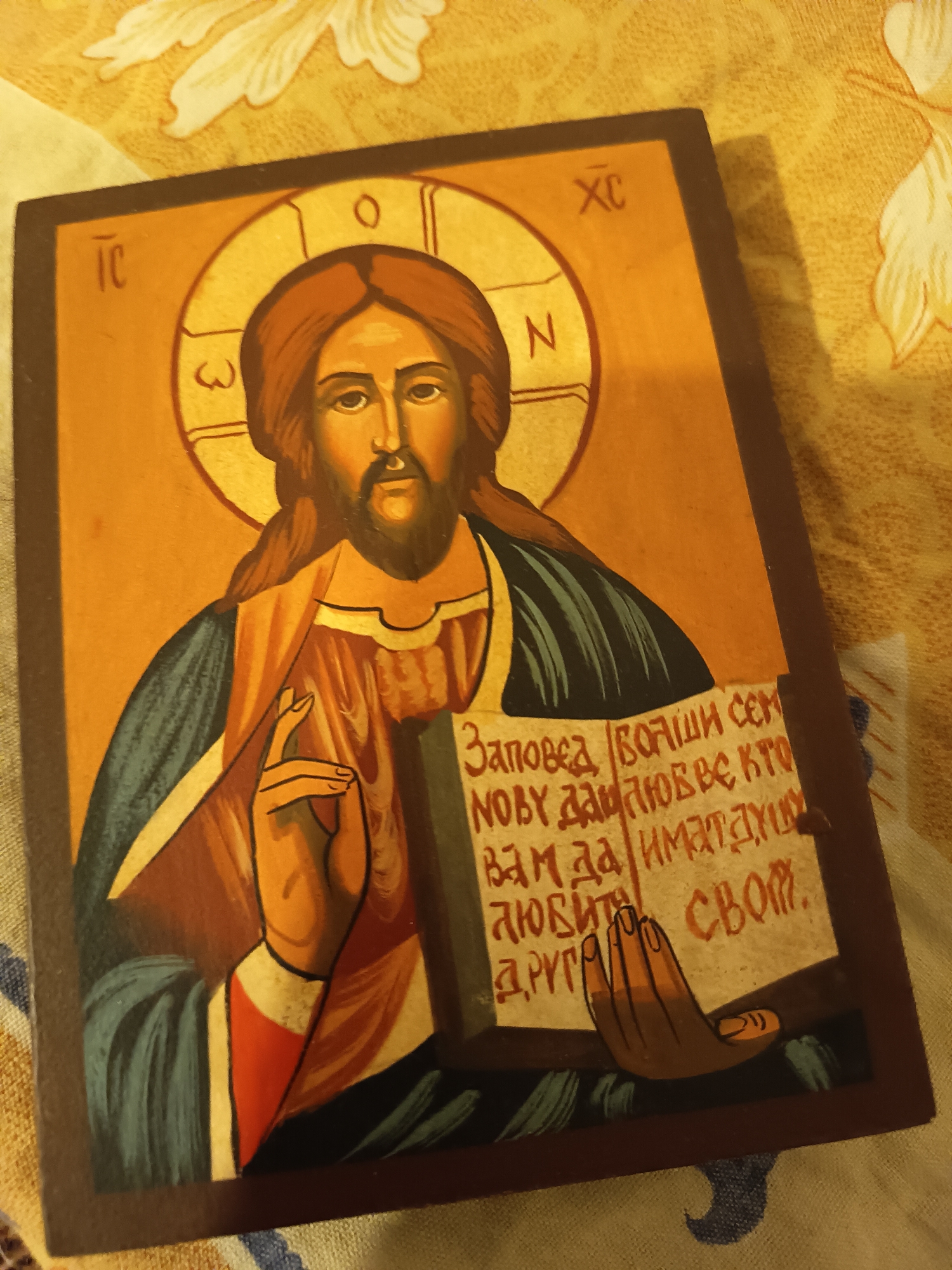 A close-up shot depicts an Orthodox Christian icon featuring Jesus Christ holding a book. The icon is framed in a dark brown wooden border and rests on a patterned fabric surface. In the upper corners are inscriptions. The main figure, Jesus, is portrayed with a dark beard and hair, and a halo behind his head. He is holding a book with Serbian Cyrillic inscriptions. His left hand is in a blessing gesture. The color palette of the icon consists of warm yellows, browns, and reds, contrasted by darker shades of blue and black. The background is blurred.
