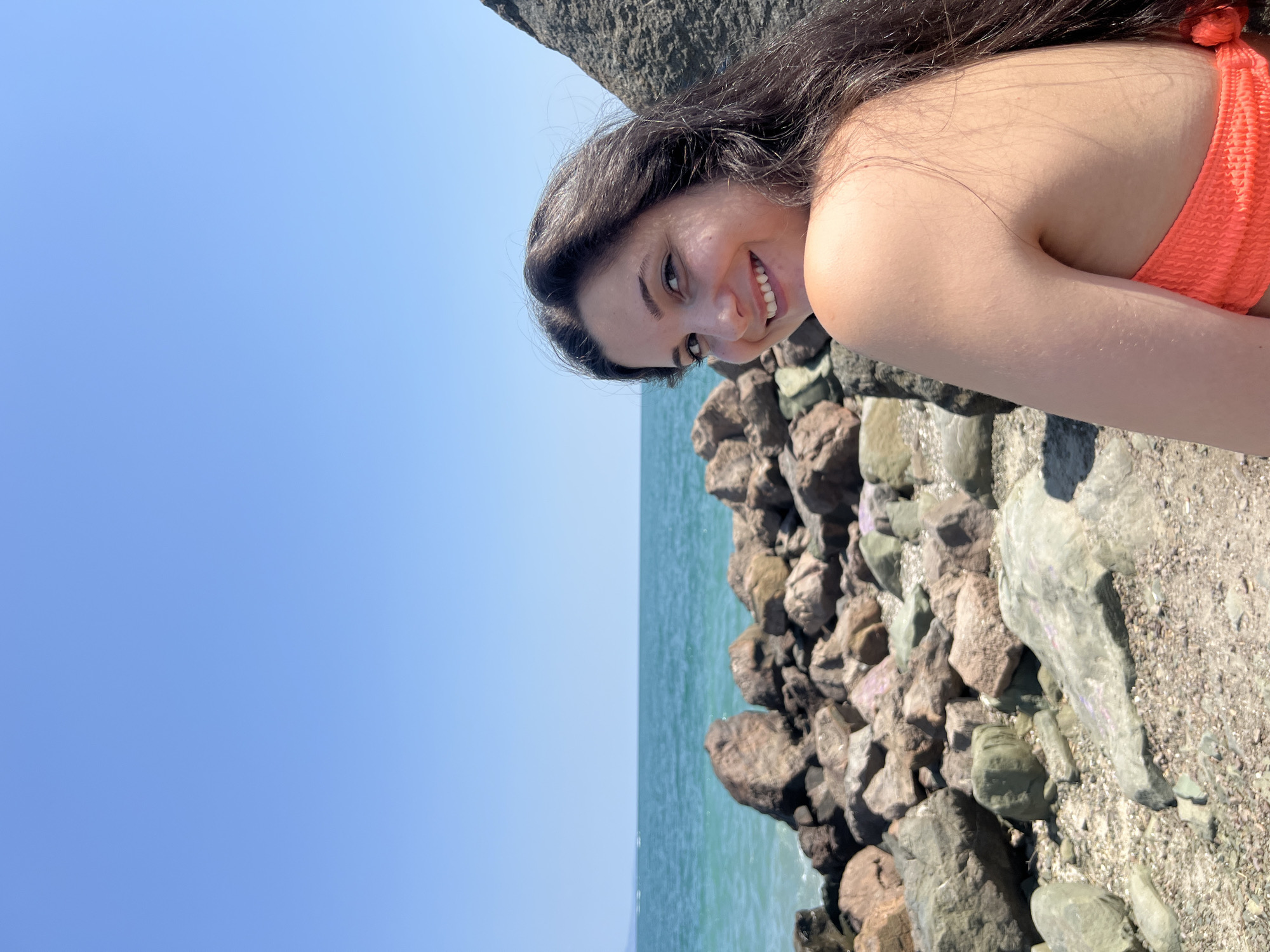A young woman with long, dark hair and a bright orange bikini top is sitting on a rocky shore in Pomorie, Bulgaria. She is looking over her shoulder at the camera with a wide smile. The sea is calm and blue behind her, with a clear blue sky above. The rocks in the foreground are a mix of brown, gray, and green, and they are scattered along the shoreline.