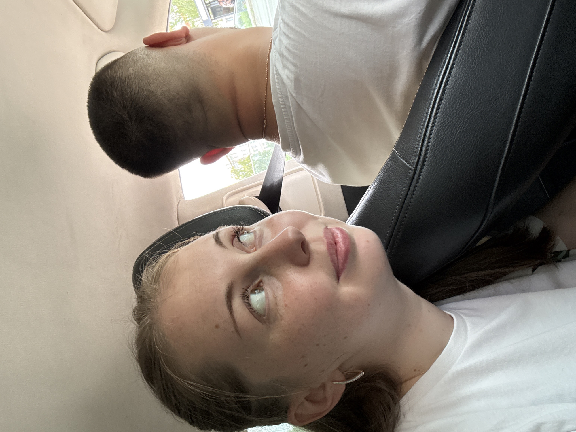 Two people are seated in the back of a car. A woman is in the foreground, looking up and to her left, with a slight smile. She has fair skin, freckles, and light brown hair pulled back. She is wearing a white t-shirt and a hoop earring. Behind her and to her right is a man, seen from the back of his head and neck. He has a buzz cut and is wearing a white t-shirt and a thin chain necklace. The car's interior is visible, with a beige headliner and a black leather seat. A seatbelt is visible across the man. Outside the car window, trees and buildings can be seen, suggesting an urban or suburban environment.