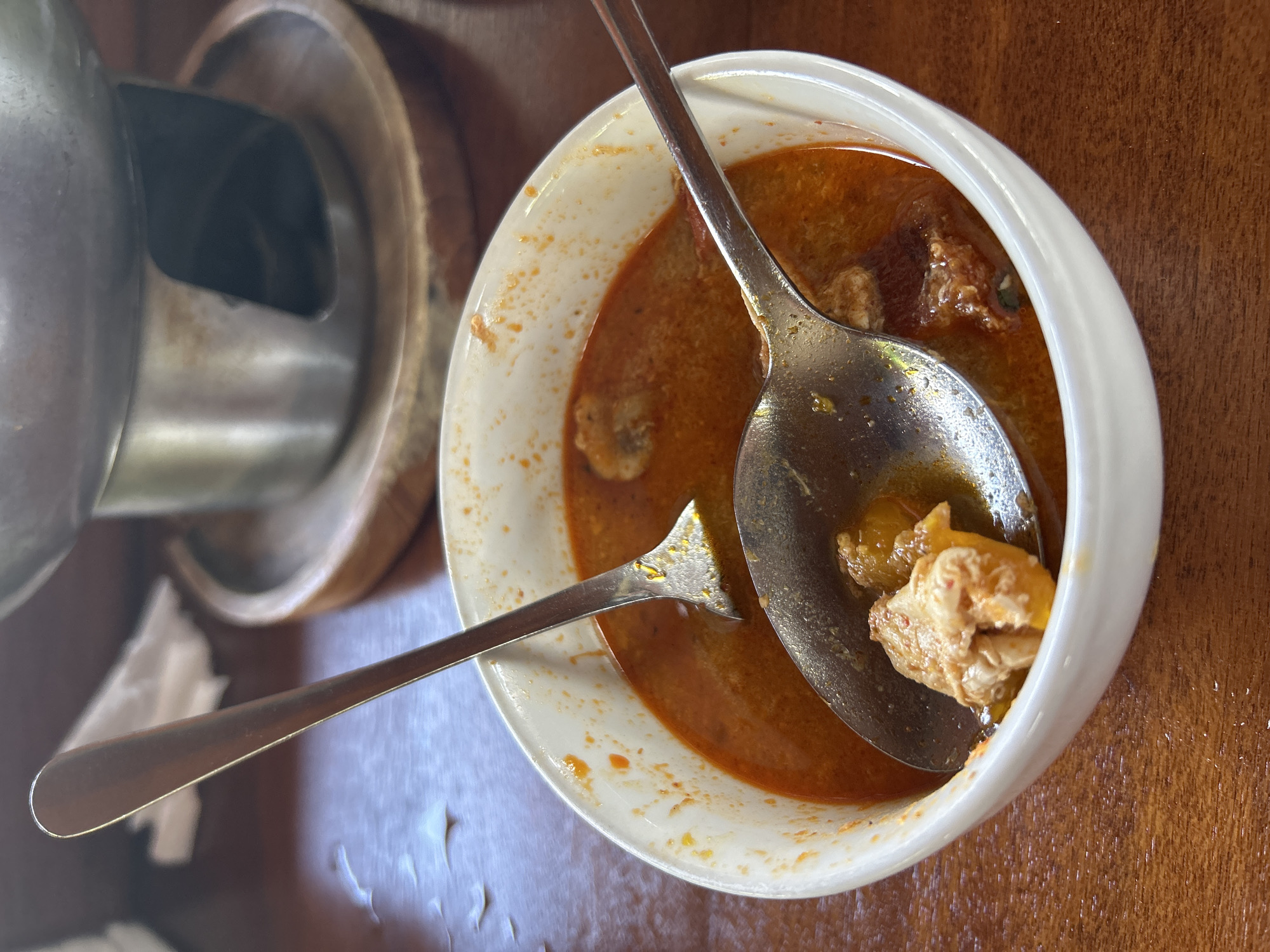 A close-up, overhead shot captures a white ceramic bowl filled with a rich, reddish-orange soup. Two metal spoons are partially submerged in the soup, one larger than the other. The larger spoon is resting at the bottom of the bowl, holding a piece of food, possibly chicken or fish. The smaller spoon is angled upwards, its handle extending out of the bowl. The soup appears to have chunks of meat and vegetables. The bowl is set on a wooden table, which shows a slight sheen, suggesting it might be polished or wet in places. In the background, a portion of a metallic chafing dish with a wooden base is visible, hinting at a meal setting. The overall scene suggests a meal in progress, likely in a restaurant or home. The location is identified as Ubud District, Indonesia.