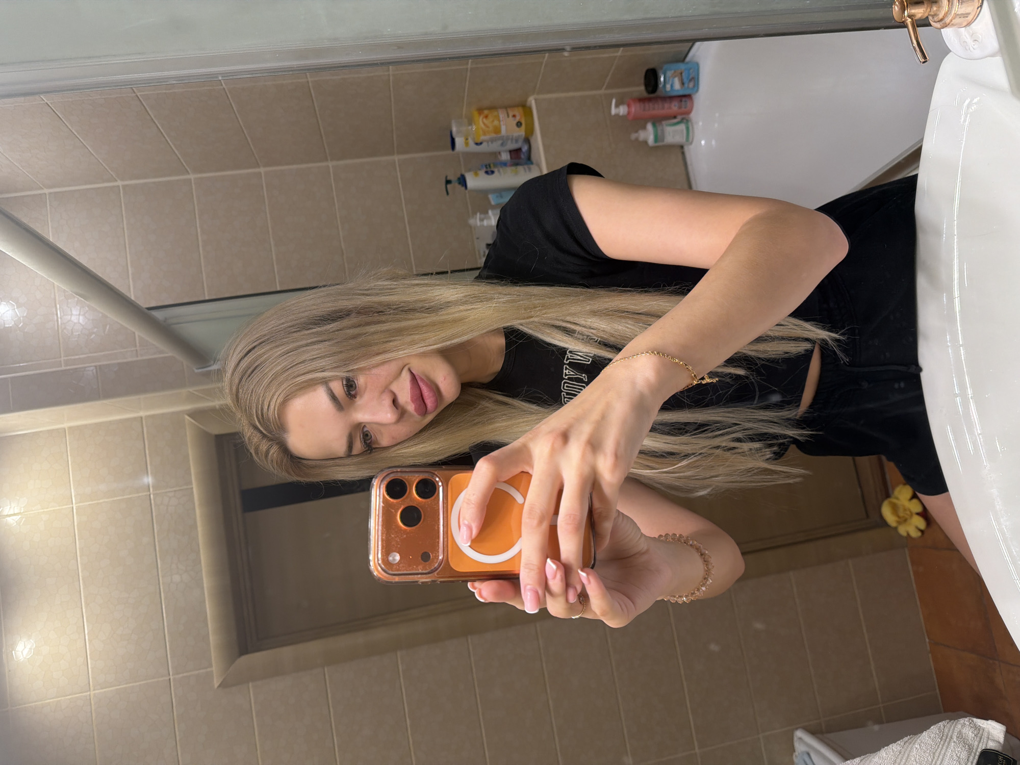 In a bathroom in Kyiv, Ukraine, a young woman with long blonde hair takes a mirror selfie. She is wearing a black crop top and black shorts. Her phone, in an orange case, is held in her hands as she poses. The bathroom features tiled walls and a bathtub with various toiletries on a shelf. A sink is visible in the foreground.