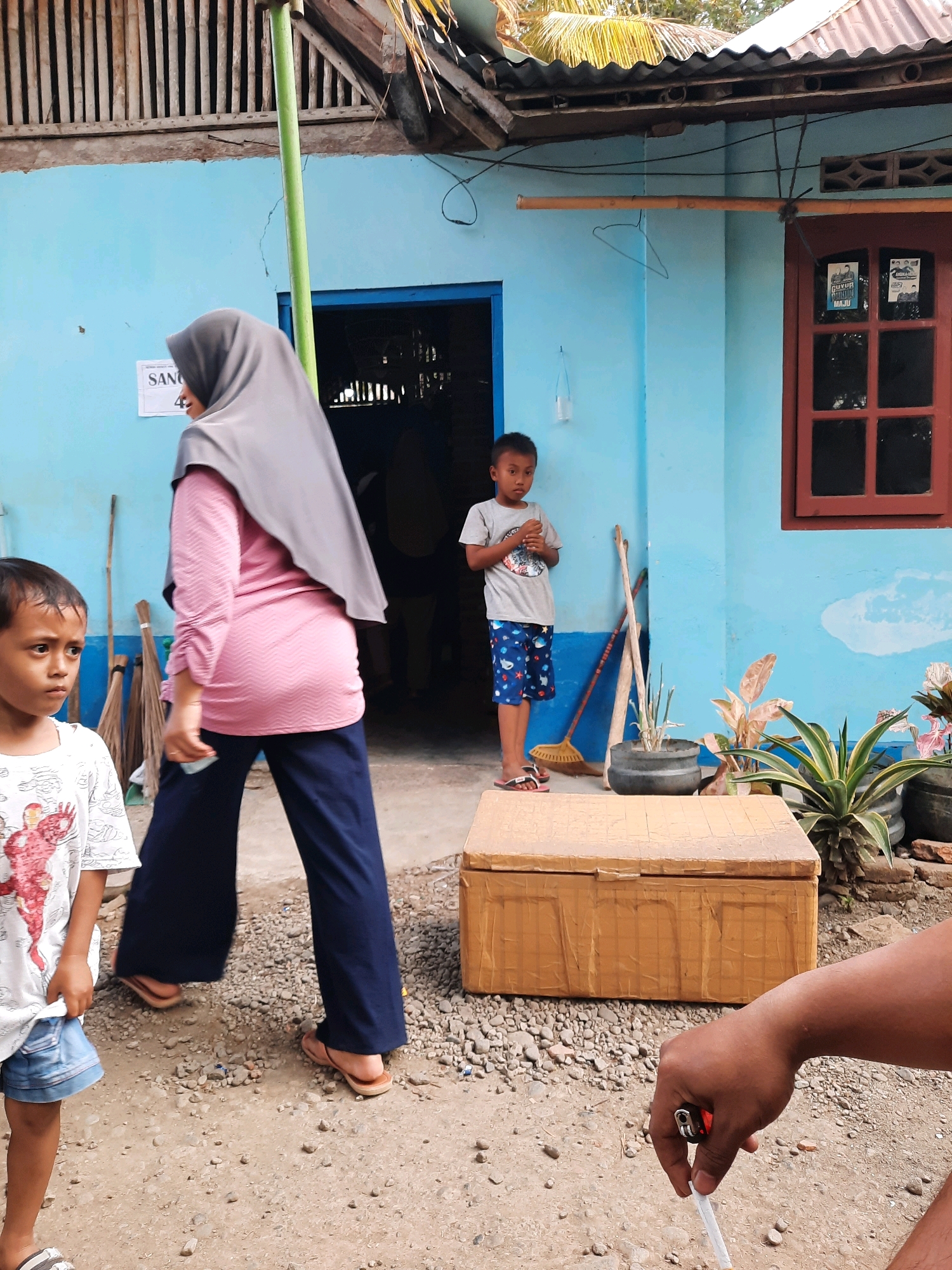 A boy stands in front of a bright blue house in Suren, Indonesia, holding a small object. A woman wearing a grey hijab and a pink and white striped shirt walks away from the viewer. Another boy, wearing a white t-shirt with a red and white design and denim shorts, stands to the left, looking towards the woman. Behind the woman, brooms lean against the blue wall. To the right of the boy in the blue house, there's a brown cardboard box on the ground, and behind it, a pot with green plants. A man's arm and hand, holding a lit cigarette and a lighter, are visible in the foreground on the right. The ground is covered in small stones and dirt. A wooden structure with vertical slats is visible above the blue house. A red-framed window is on the right side of the house.