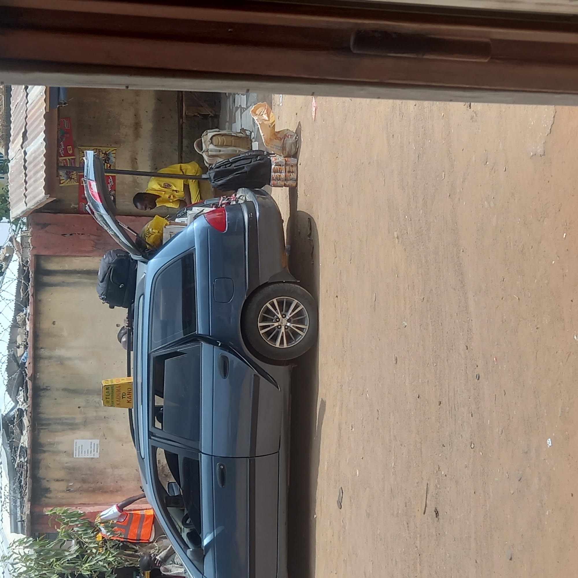 The image depicts a blue station wagon parked on a dirt road in Dadi Riba, Nigeria. The rear hatch of the car is open, revealing the interior. A man wearing a yellow shirt is near the rear of the car. Luggage, including a backpack and several smaller items are visible in the opened back of the car. Another person wearing a white shirt and an orange vest is near the front of the car. A sign is visible on the side of the car that says "RTLAN (Ride Sharing) KADUNA TO KANO". The surroundings show buildings with corrugated metal roofs and barbed wire fences. The ground is dusty.