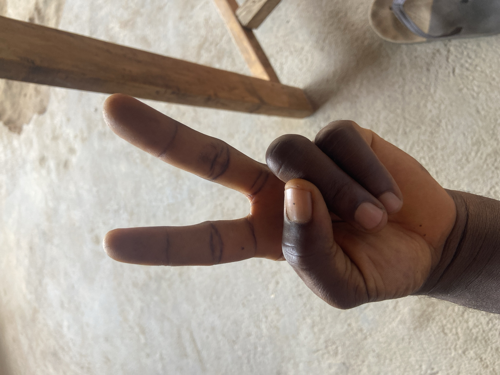 A close-up shot displays a hand making a peace sign. The hand is darker-skinned. The fingers are extended in a "V" shape, while the other fingers are curled inwards. The backdrop shows a light-colored surface. There are wooden planks and a flip-flop in the background. The image was taken in Mararaba, Nigeria.