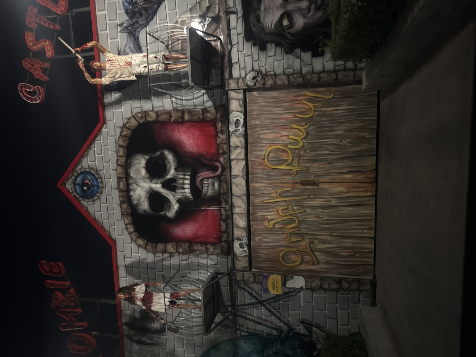 The image displays the illuminated facade of a horror-themed attraction at night. The structure is designed to resemble a grey brick castle, featuring red trim along the roofline and a central circular decoration with a blue eye. Above the building, partially visible red neon signs spell out "ZOMBIE" on the left and "CASTLE" on the right.
Two zombie figures are positioned on metal balconies: a female figure in a blood-stained white dress stands on the left, and a male figure in a blood-stained white shirt and pants, holding a white club, stands on the right. Between these figures, a large arched opening frames a painted skull with an extended tongue against a red background. Below this arch, two small white skull decorations are affixed to the brickwork.
To the left, below the female zombie, a purple and yellow sign is partially visible on the brick wall, displaying "1000Դ" along with Armenian text, indicating a price of 1000 Armenian Dram. The main entrance consists of two tall, dark wooden-textured doors. Golden-yellow stylized Armenian script is painted across these doors, reading "ԶՈՄԲԻ ԴՂՅԱԿ," which translates to "ZOMBIE CASTLE."
The ground directly in front of the entrance is a dark, paved surface. A small green bush is visible at the lower right base of the building. The overall scene suggests an amusement park or entertainment venue in Yerevan, Armenia, given the specific Armenian language and currency visible. No live individuals are present; all figures are static props.
