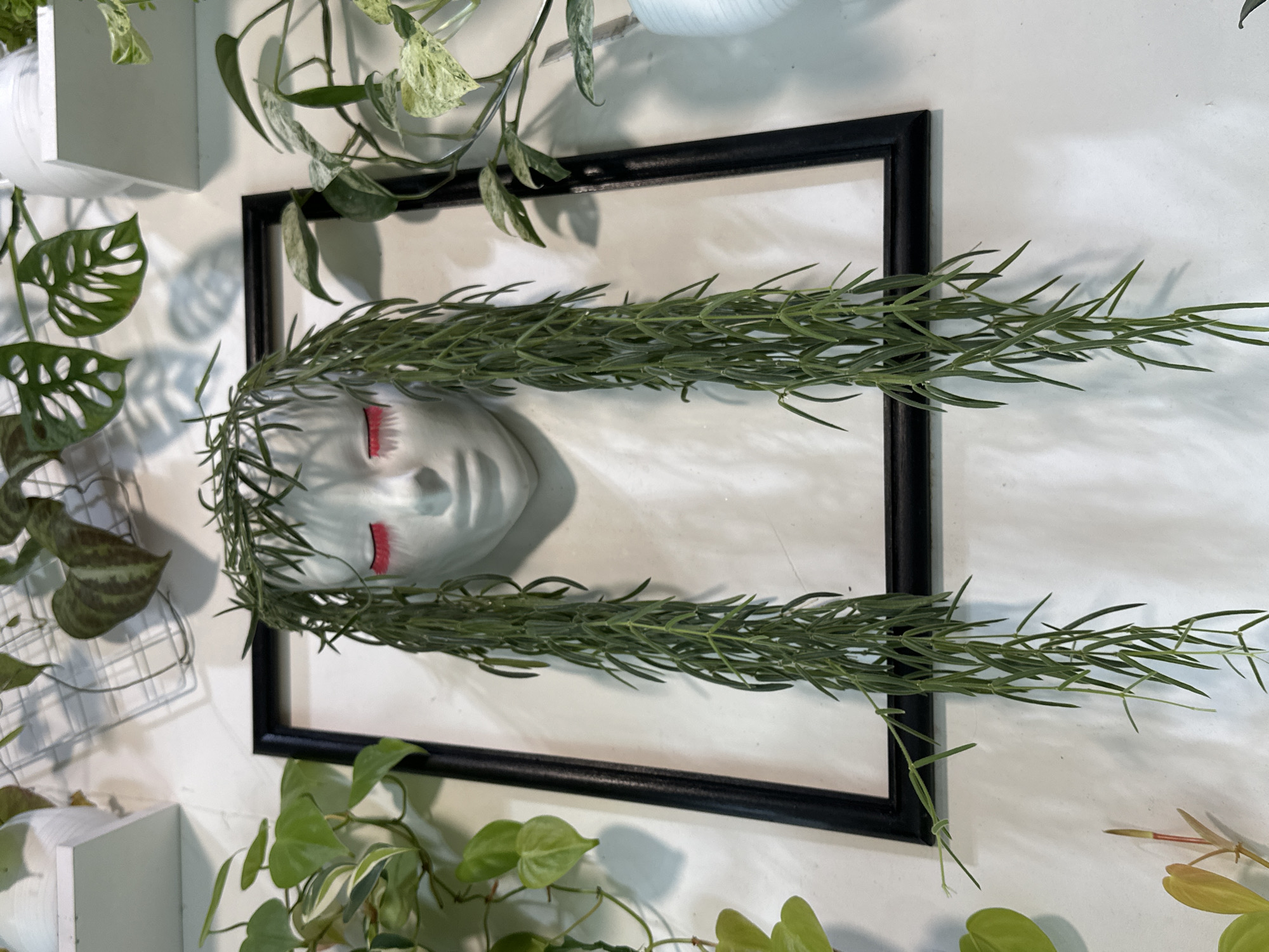 A wall decoration features a white, face-shaped planter with a dark frame behind it. The planter has closed eyes with bright pink eyelashes and long, green, trailing plant stems that resemble hair. Various green plants are visible around the planter, some with heart-shaped leaves and others with variegated patterns. The background is a textured white wall, and a white shelf is visible in the upper left corner. The scene appears to be indoors, possibly in a home or a shop specializing in plants or home decor. There is no indication of this scene being in Moscow, Russia.