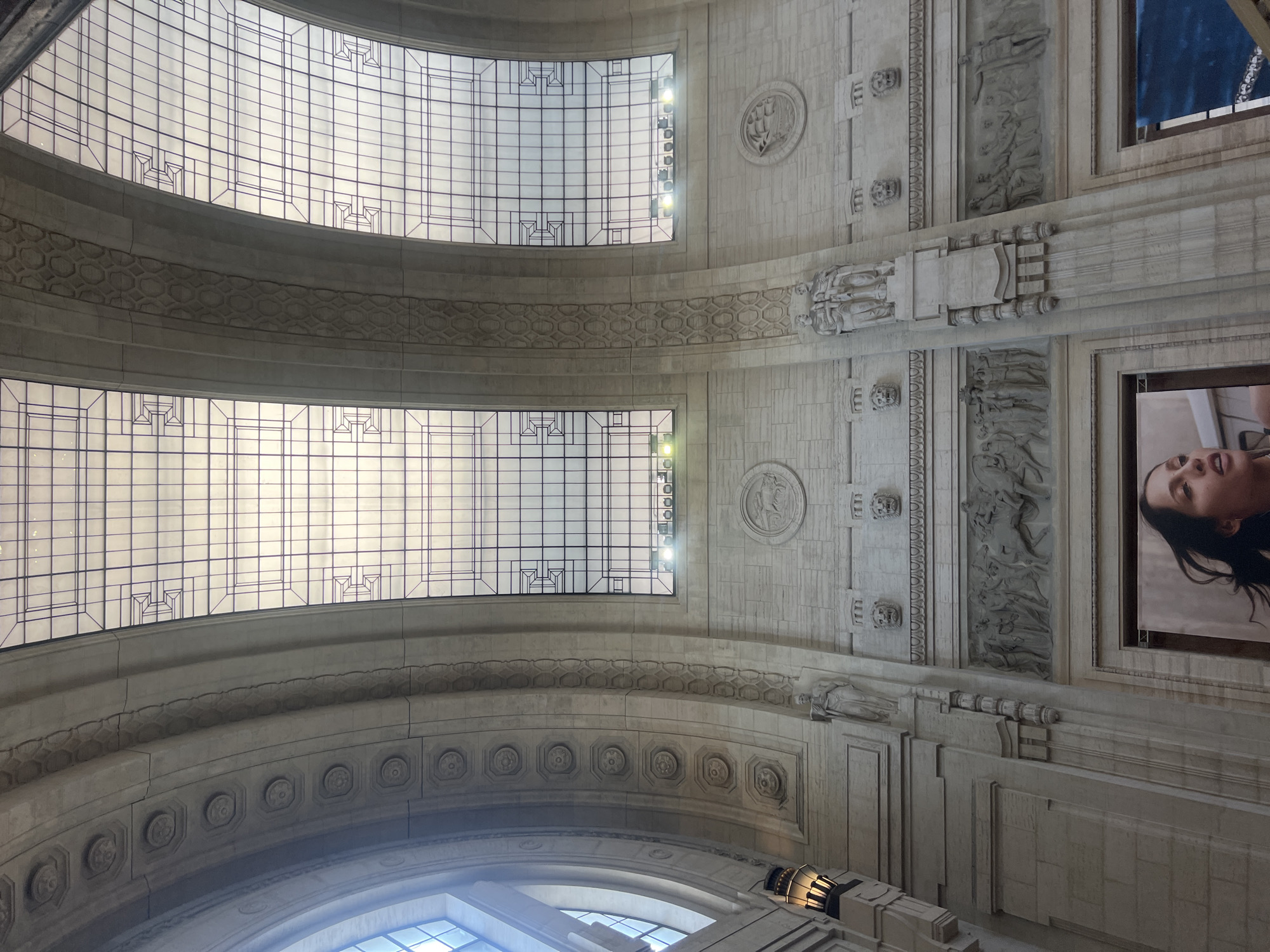 The image depicts the interior of Milan Central Station, Italy, looking upward at the ceiling. The architectural features include arched windows with geometric glass patterns, separated by ornamental stone arches with repeating patterns. The walls have decorative relief sculptures, medallions, and architectural details. A modern photograph hangs on the right wall, featuring a woman's face. The perspective is looking upwards, emphasizing the height and detail of the station's ceiling. The dominant colors are gray and white, reflecting the stone construction.