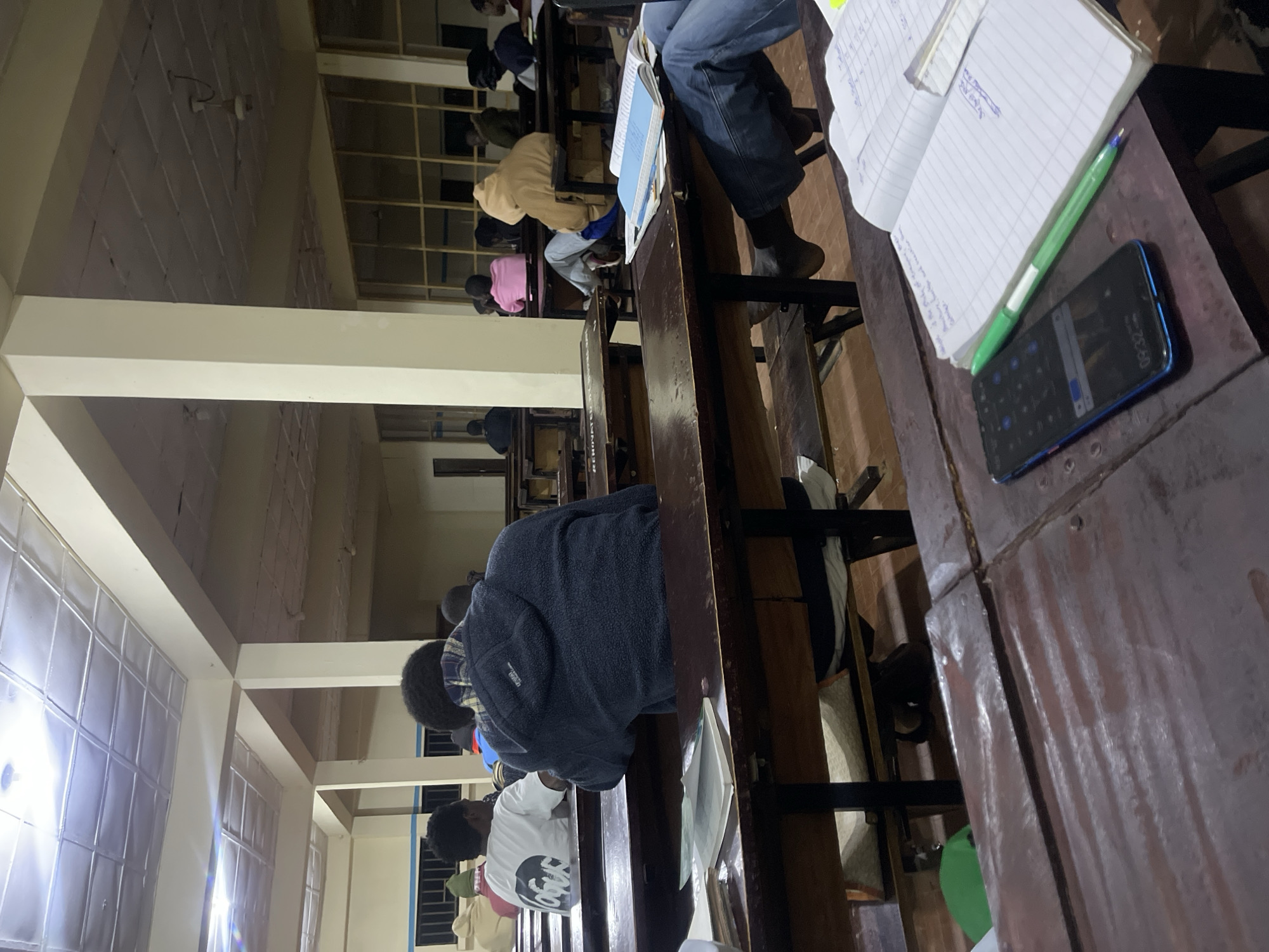 The image depicts an indoor scene, likely a classroom or study hall, with approximately ten individuals seated at wooden desks. The room is lit by overhead fluorescent lights, and a ceiling fan is visible. The architecture suggests a functional, perhaps institutional, building with visible support beams and paneled ceilings. The desks are old and worn, with some appearing to be attached to benches.
In the foreground, on a desk, a smartphone displaying the time "09:32" is placed next to an open notebook with handwritten notes and a green pen. The notebook is open to a page with a ledger-like layout.
People are mostly facing away from the camera, engaged in studying or listening. One individual in the middle ground wears a dark blue hooded jacket. To their left, another person in a white t-shirt with a graphic print is visible. In the background, other students are seated at desks, some wearing hoodies in various colors, including pink and tan. The overall atmosphere appears to be one of quiet study or instruction.
The context of Rafin Par, Nigeria, suggests this is likely a school or educational facility within that region. The image captures a moment in an academic setting, focusing on the individual study environment and the general classroom setup.