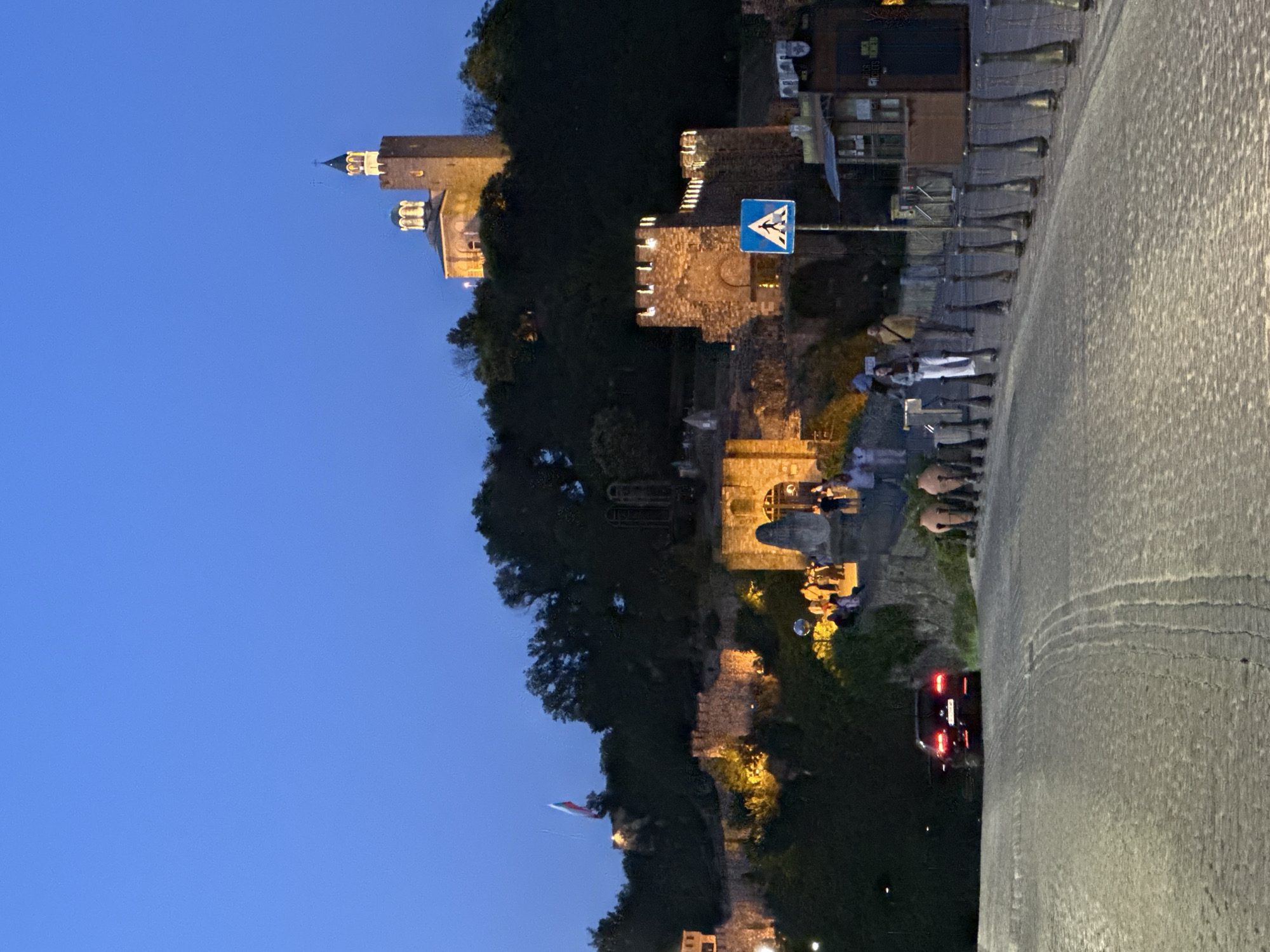 The image shows Tsarevets Fortress in Veliko Tarnovo, Bulgaria, at dusk. The fortress is illuminated, and the Bulgarian flag is visible on a flagpole. A cobblestone road leads to the fortress, with several people walking and standing around. A black car is parked on the side of the road, and there are streetlights visible. The sky is a deep blue, indicating the transition from day to night.