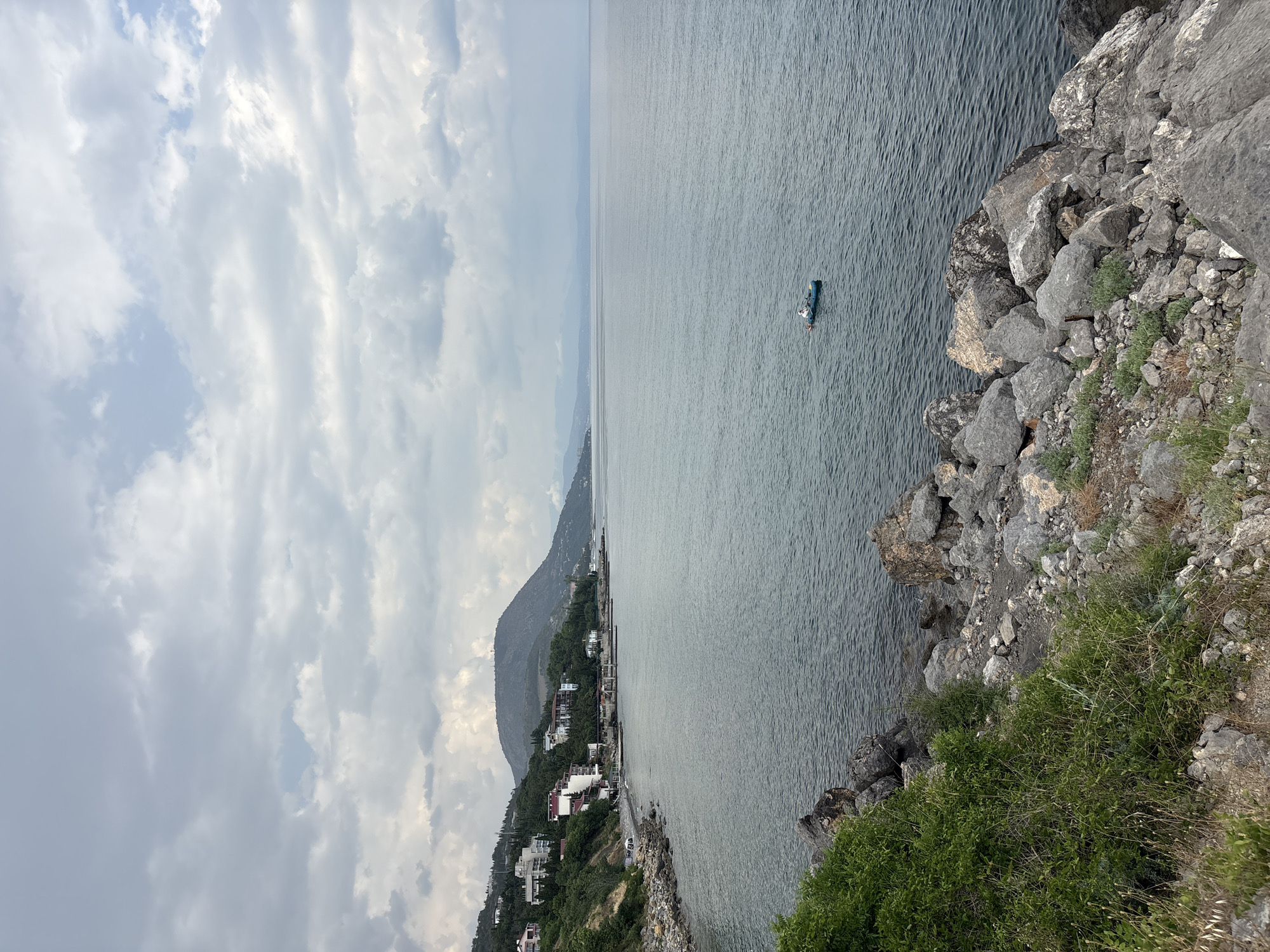 The image depicts a coastal scene with a body of water, a shoreline, and a hillside with buildings. The water is a calm, dark blue, with gentle ripples. A small, blue boat with a single person in it is visible in the middle of the water, rowing. The shoreline is rocky and uneven, with some sparse vegetation growing amongst the rocks. On the hillside overlooking the water, there are several buildings, appearing to be houses or small hotels, with white and red facades. The hillside is covered in lush green trees and vegetation. In the background, a large, rounded mountain rises from the landscape, its peak shrouded in a light haze. The sky is overcast with a mix of light and dark grey clouds, with patches of pale blue visible. The overall atmosphere is serene and slightly moody due to the cloudy sky. This scene is located in the city of Bondarenkovo, Ukraine.