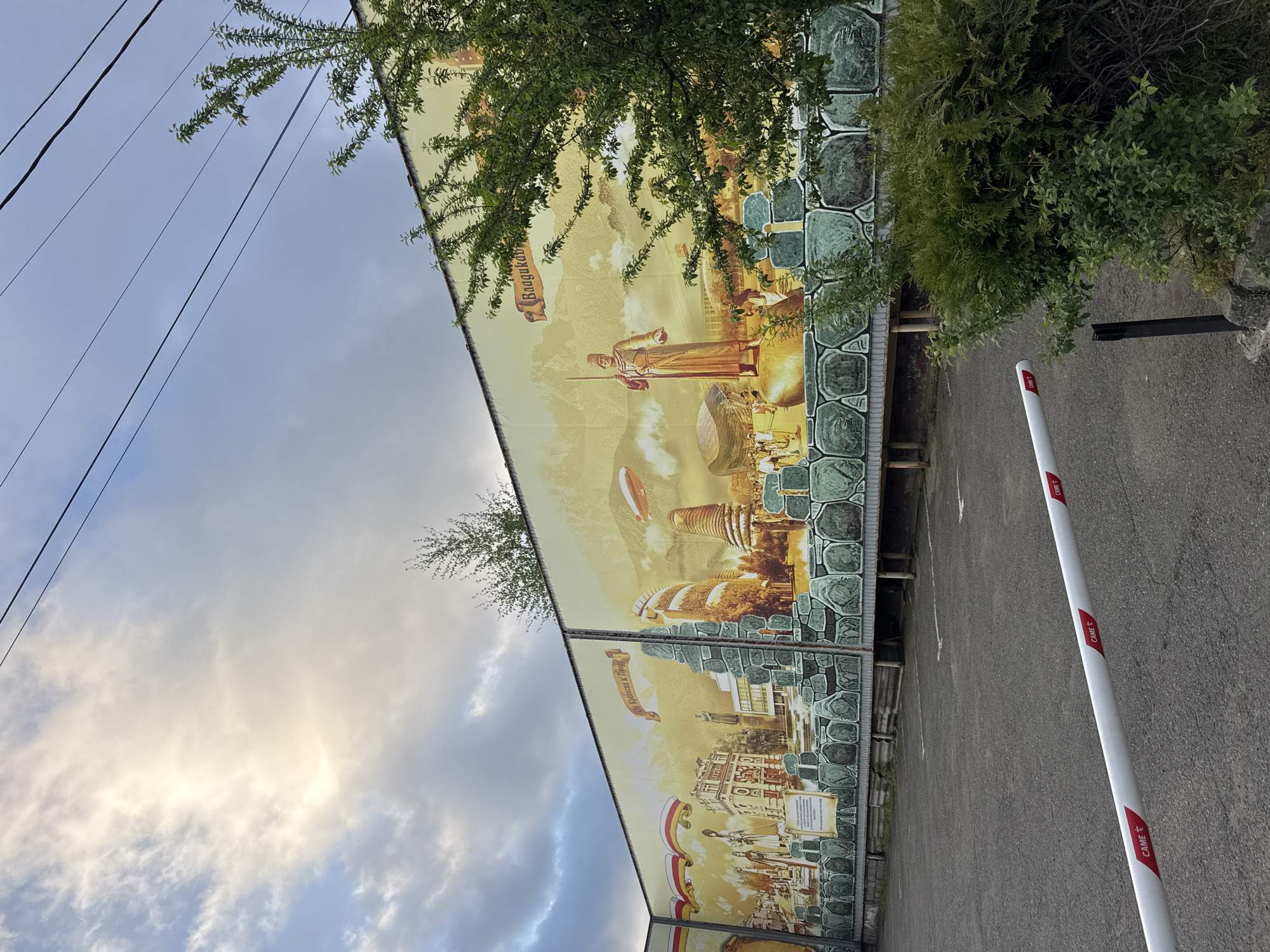 The image depicts a large mural on a building in Akhieli, Georgia. The mural features scenes of the city, including historical figures, architecture, and landmarks, set against a backdrop of mountains. The mural is bordered with a stone-like pattern. Overhead power lines are visible against a cloudy sky. A gate arm with "CAME" logos is located on the right side of the image in front of an asphalt parking lot. Green foliage partially obscures the mural.