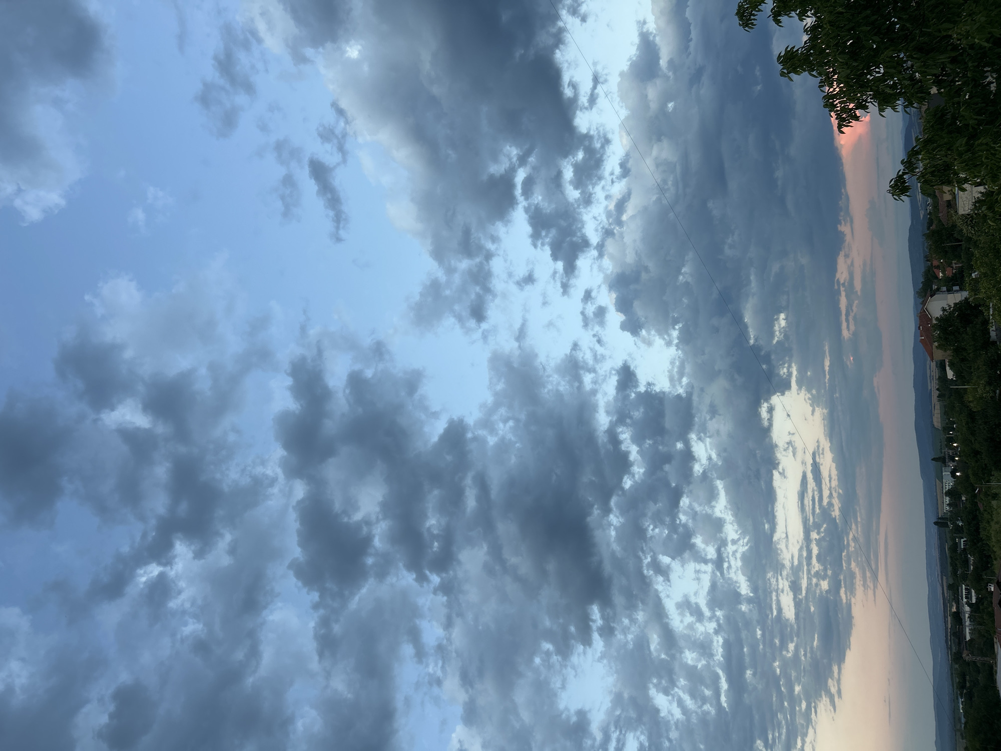 The image is a vertical, medium-shot photograph of a cloudy sky over a distant landscape of buildings and trees. The sky occupies approximately two-thirds of the frame and is filled with a mix of dark gray and lighter blue clouds. Some of the clouds are large and dense, while others are wispy and scattered. The lower third of the image shows a panoramic view of a town or village nestled in a valley. Buildings with red tile roofs are visible, as are numerous green trees. In the far distance, a hazy line of hills or mountains forms the horizon. A single thin, dark wire is visible diagonally across the sky from the top left to the bottom right. On the right side of the frame, the leaves of a dark green tree are visible in the foreground, partially obscuring the sky and landscape. The lighting suggests it is either early morning or late evening, with a subtle pinkish hue near the horizon, possibly indicating the presence of a sunset or sunrise.