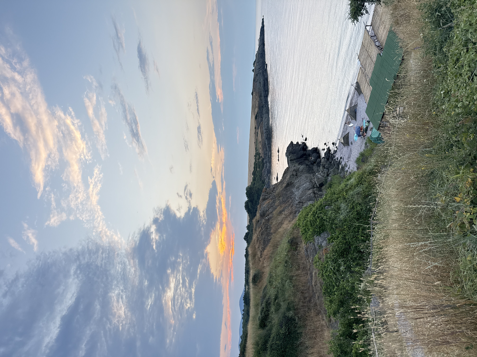 This image captures a coastal landscape near Varvara, Bulgaria, during what appears to be sunset or sunrise. The sky is a blend of soft blues, grays, and oranges, with scattered clouds. The sea is calm and reflects the sky's colors.
In the foreground, a grassy, overgrown hillside slopes down towards the sea. The vegetation consists of dry grasses and green bushes. A rustic wooden fence is visible, partially obscured by the tall grasses.
Further down the slope, a small beach is nestled between rocky outcrops. Several conical tents, typical of camping or glamping setups, are pitched on the pebble beach. A few beach umbrellas and people are visible near the tents. Adjacent to the beach, a small cluster of wooden structures with green and thatched roofs are situated, suggesting some form of amenity for beachgoers, possibly a cafe or accommodation.
The coastline is characterized by rugged, rocky cliffs covered in patches of green vegetation. The sea meets the shore along a rocky edge and a small pebble beach area. In the distance, a hilly landscape with scattered trees forms the horizon. The overall scene conveys a sense of tranquil, natural beauty at the edge of the sea.