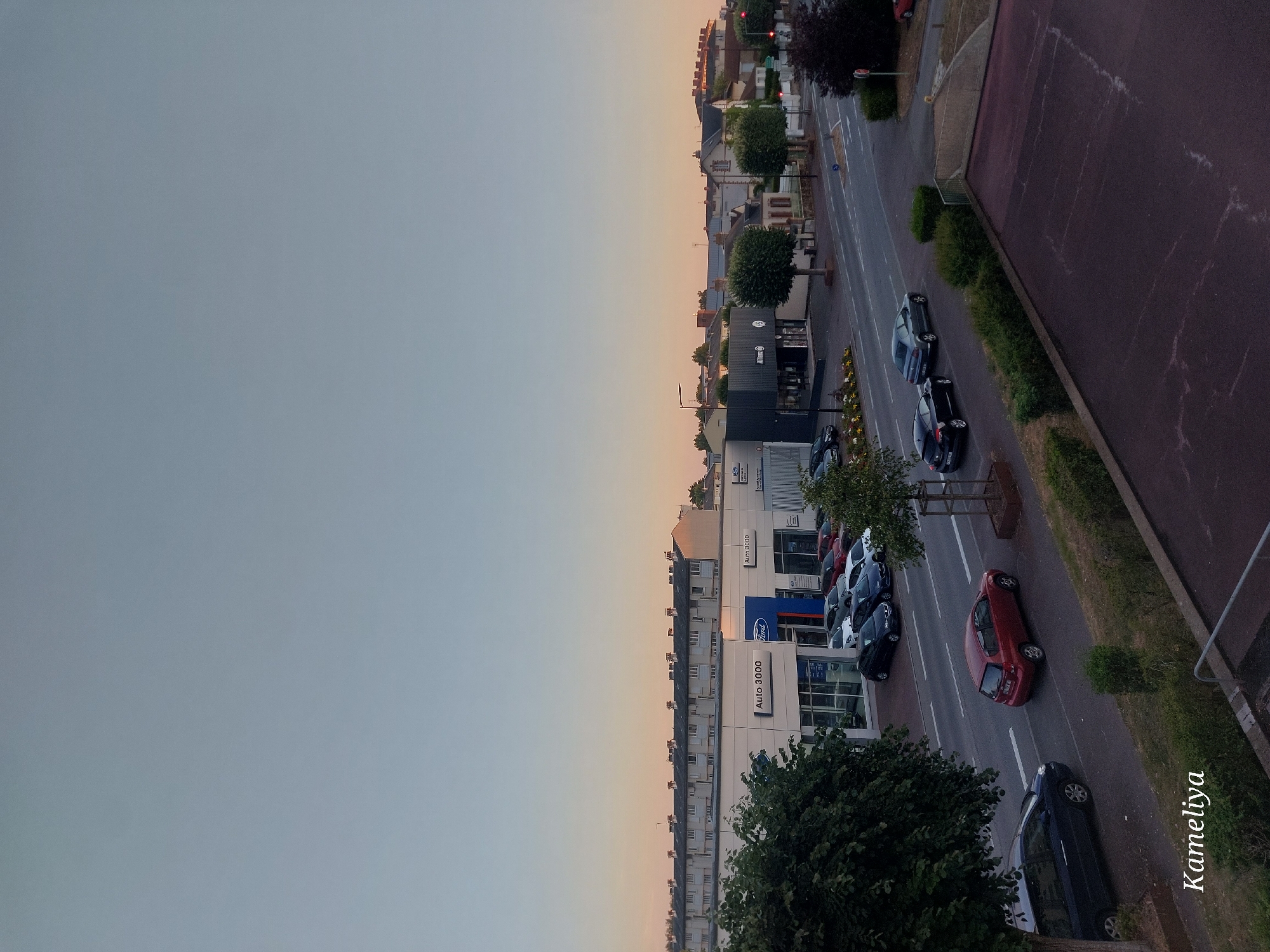 The image is a high-angle shot of a street scene in Alençon, France, likely captured during the late afternoon or early evening, given the soft, warm light on the horizon. The sky is a gradient of pale blue and soft orange.
In the foreground, a dark reddish-purple paved surface with some white markings takes up the bottom right portion of the frame. To the left of this surface, a green grassy area with scattered bushes leads to a road.
The road runs diagonally across the middle of the image. Several cars are visible on the road and parked in front of businesses. On the left side of the road, a dark blue sedan is driving away from the camera. Further down the road, a red hatchback is also driving away. Across the road, a dark sedan and a silver sedan are parked. Beyond them, there's a row of parked cars, including a dark blue hatchback, a white sedan, and a dark blue sedan.
On the left side of the road, there is a row of buildings. One building prominently displays "Auto 3000" and a "Ford" logo, suggesting a car dealership. Adjacent to it, another building is visible with "Auto 3000" signage and a dark facade. Further down the street, other businesses with signs like "Allianz" are visible, along with trees and residential buildings in the background. A large tree with dark green foliage is present on the far left of the image.
The overall environment appears to be a commercial area with residential buildings in the background, situated in a town or city. The presence of streetlights and the layout of the buildings indicate an urban or suburban setting. The watermark "Kameliya" is visible in the bottom left corner.