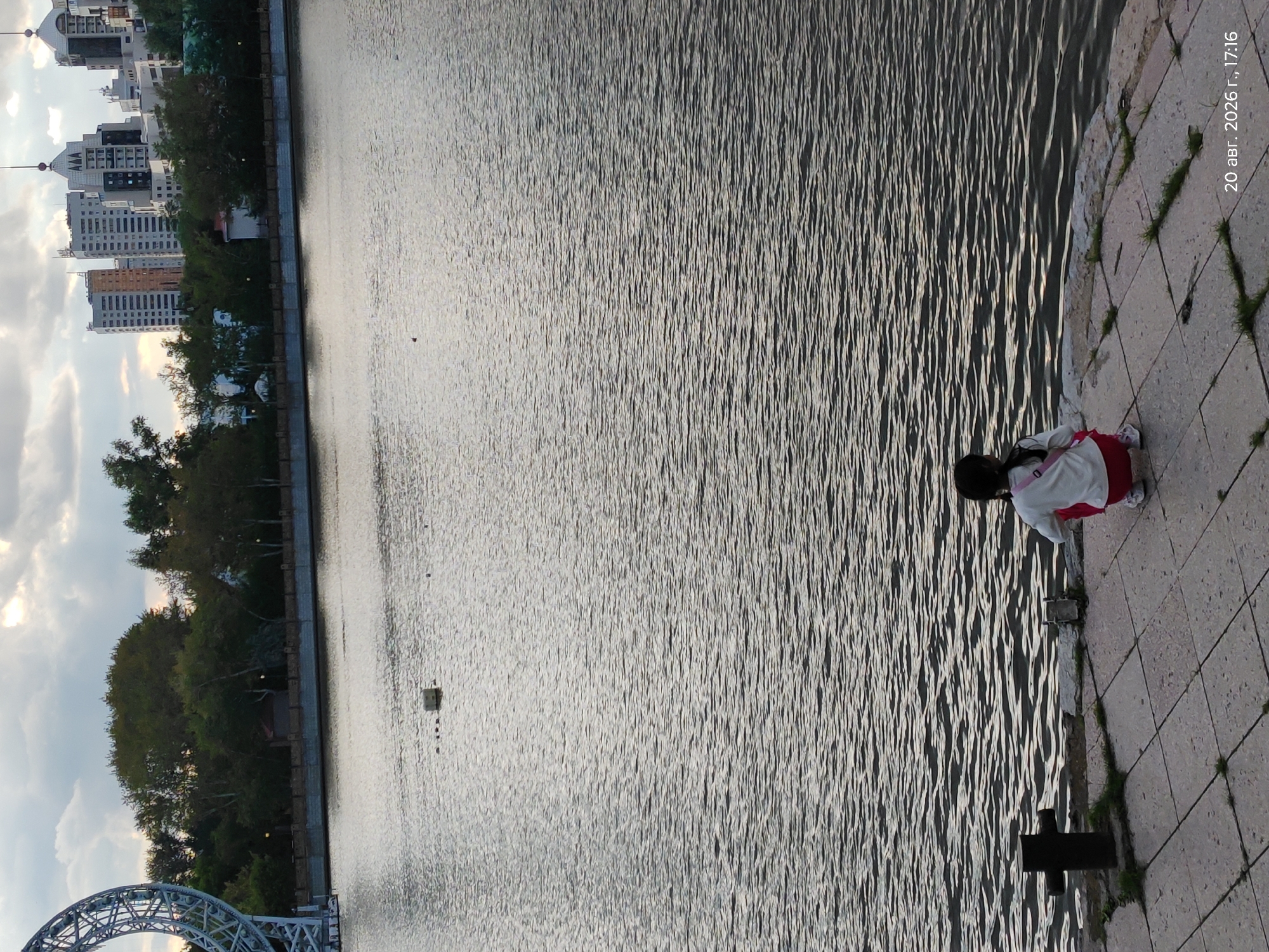 The image captures a scene from the city of Astana, Kazakhstan, likely along the Ishim River embankment. In the foreground, a young child, viewed from behind, is crouched down on a paved walkway overlooking the water. The child wears a white long-sleeved shirt and red pants, with a pink bag slung over their shoulder. The walkway is made of large, square stone tiles, with patches of grass growing between them. A black bollard stands to the left of the child. The water in the river is rippled, reflecting the overcast sky. In the background, a line of trees separates the riverbank from a cluster of modern high-rise buildings characteristic of Astana's cityscape. To the far left, a portion of a large, circular metallic structure is visible, possibly part of a Ferris wheel or other urban landmark. The overall atmosphere is serene, with a touch of urban ambiance.