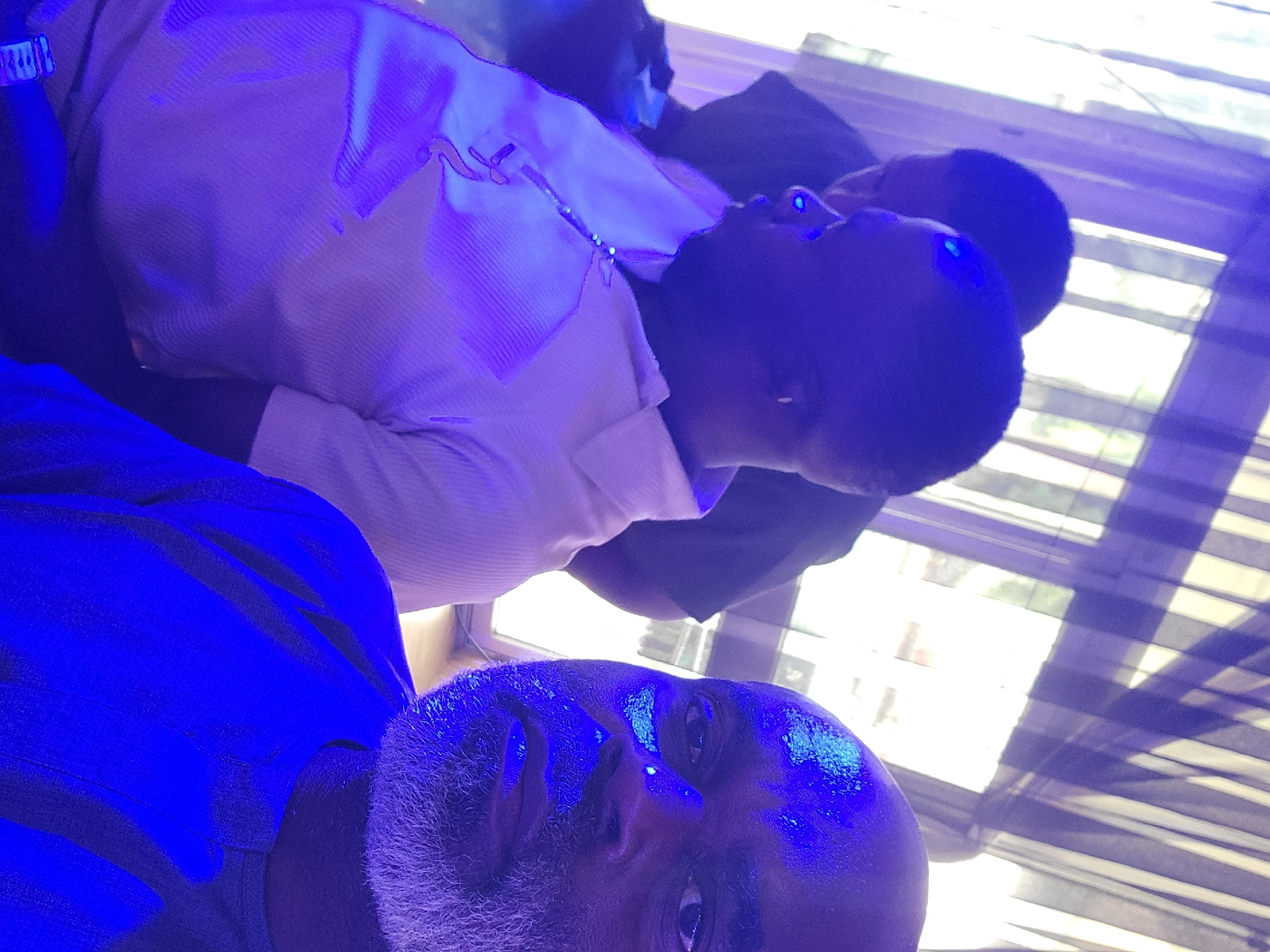 A close-up, high-angle shot depicts at least three individuals indoors, under strong blue lighting. The person closest to the camera is an older man with gray facial hair, whose face fills the bottom portion of the frame. Behind him, slightly out of focus, is a younger man wearing a light-colored shirt. Further back, a third person with short dark hair is visible. The background appears to be a window with blinds or shutters. The location in Barden Yana, Nigeria, cannot be determined from the visual information alone.