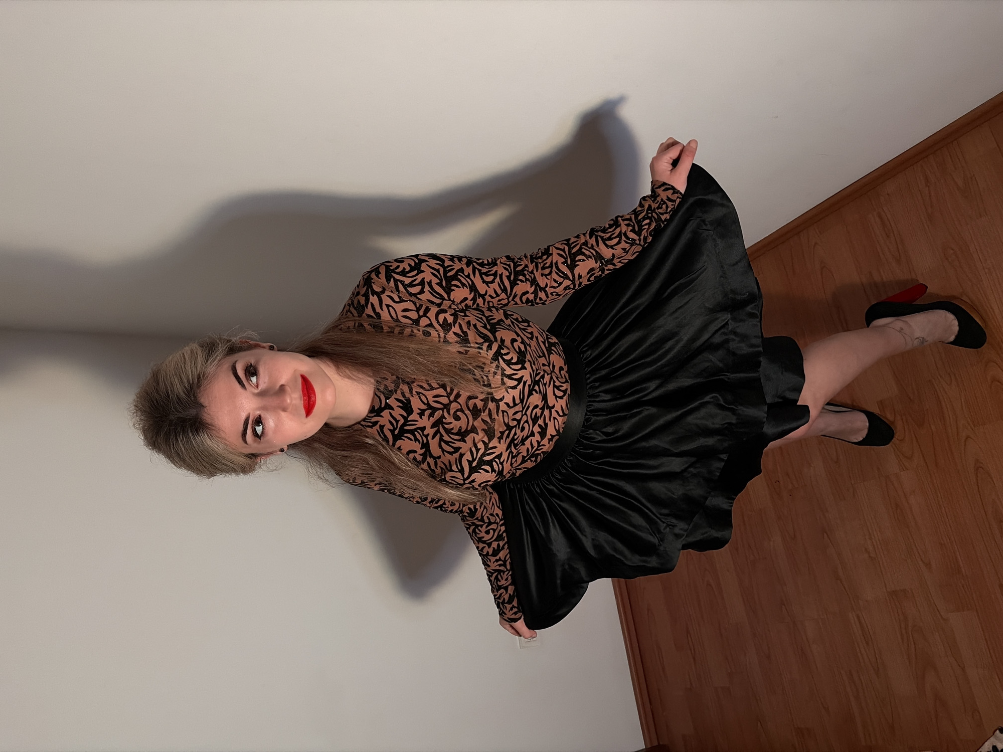 In Budva, Montenegro, a female subject stands indoors, positioned in a corner formed by two plain, light-colored walls. The floor is composed of light-brown wooden planks with a narrow baseboard along the wall. The subject has light blonde hair, partially styled up, and is wearing bright red lipstick. She is attired in a long-sleeved top featuring a light brown base with black abstract patterns, and a voluminous, shiny black skirt. She wears black high-heeled shoes, with the right shoe distinctly displaying a red heel and a light-colored anklet on her right ankle. The subject is looking towards her left, with a slight smile, holding the sides of her skirt with both hands. Illumination from an elevated left source casts a pronounced silhouette of the subject on the wall behind her. A white electrical outlet is visible on the wall to her left.