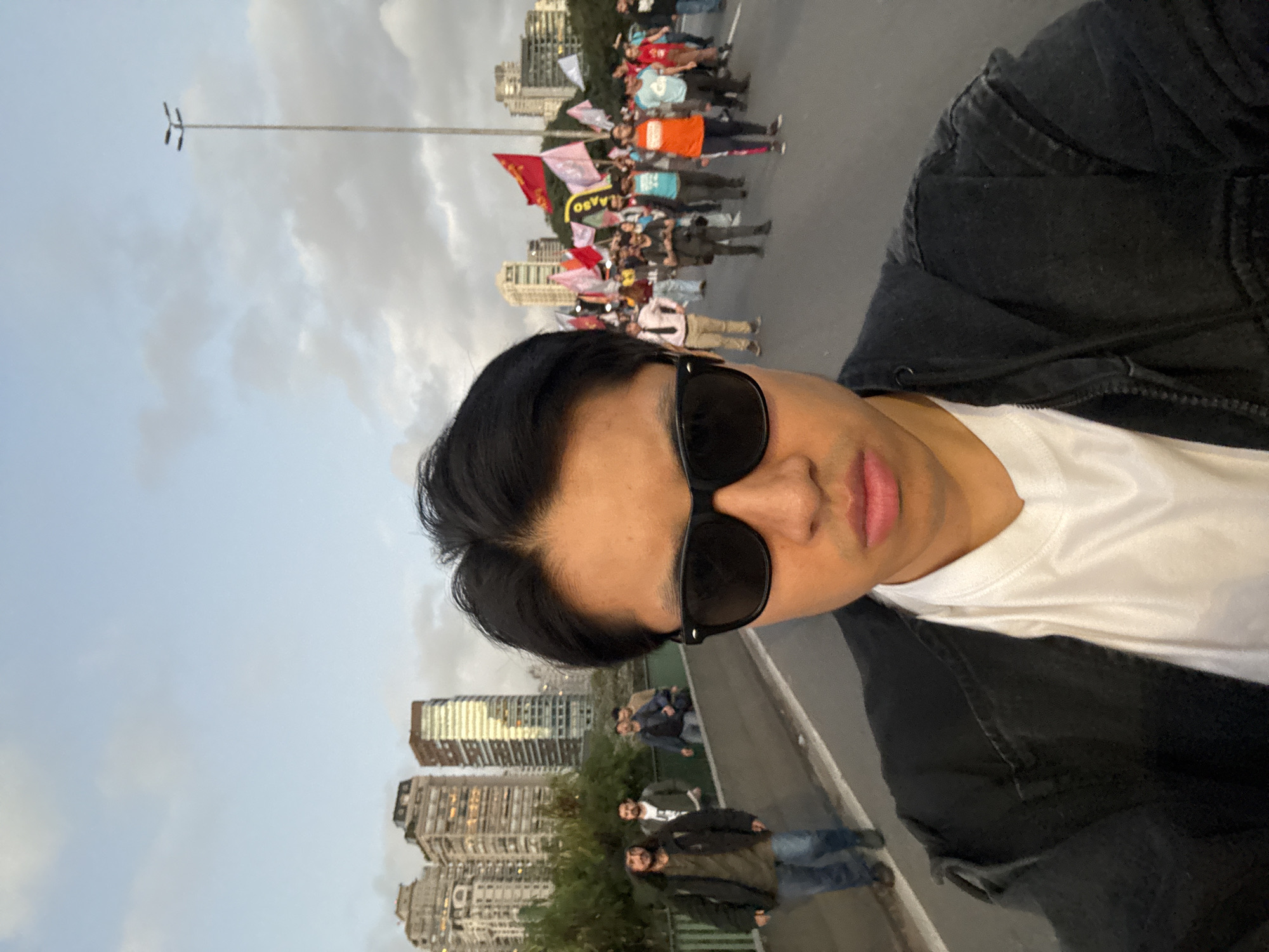 A first-person perspective photograph captures a person wearing sunglasses, a white t-shirt, and a black jacket. The background reveals an urban scene with tall buildings characteristic of the city of São Paulo, Brazil. A group of people are gathered, possibly protesting or demonstrating, as they are holding flags and banners. A tall metal light pole stands in the distance under a cloudy sky. Additional people appear to be walking on a sidewalk or pathway along a bridge or elevated walkway.