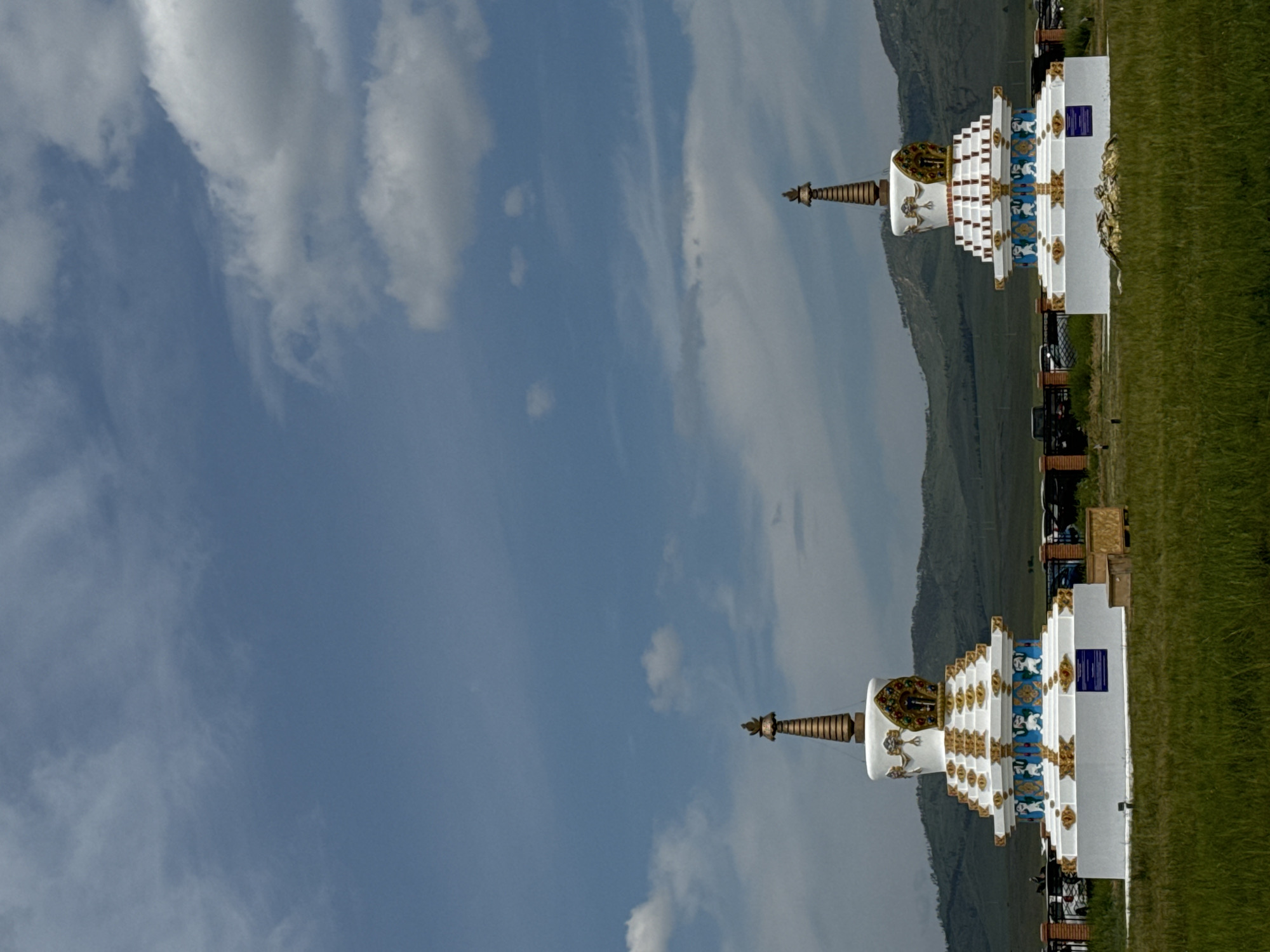 Two ornate stupas stand in a grassy field under a blue sky with scattered clouds. The stupas are white with gold and blue decorative elements. The stupas appear to be identical, with multiple tiered layers and intricate designs. In the background, a line of green hills stretches across the horizon. The ground in the foreground is covered with green grass, and there is a fence with some vehicles behind it. The location is the city of Orongoi, Russia.