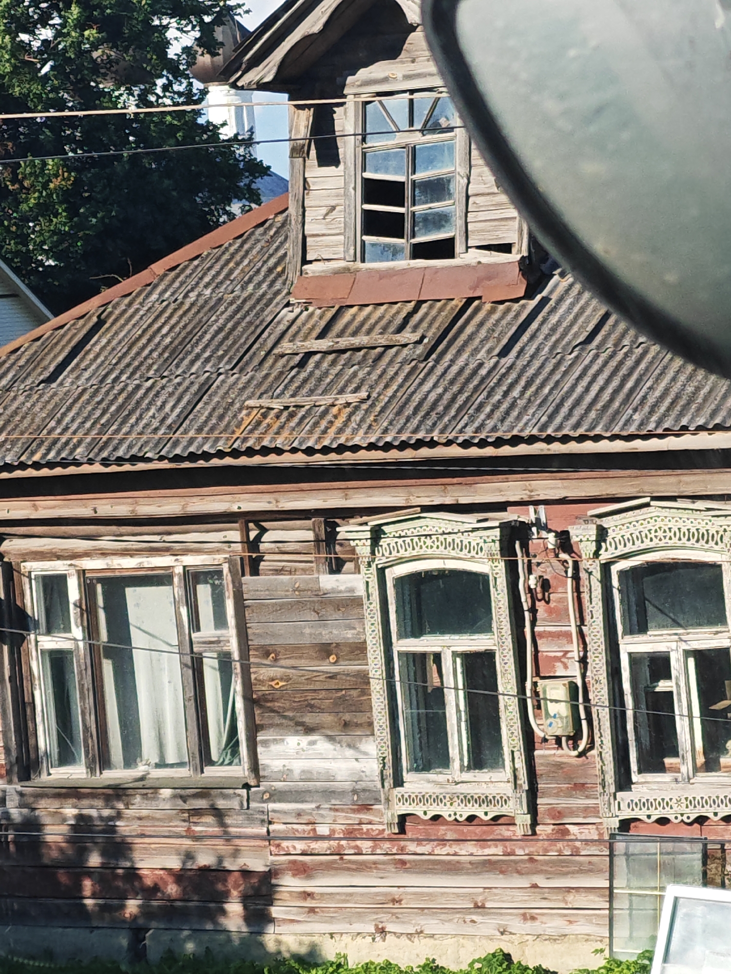A weathered, wooden, two-story house is pictured in Zavidovo, Russia. The house features a corrugated metal roof, and several windows. The lower floor has two windows with decorative wooden trim. The upper floor has a dormer window with broken panes. The house appears to be in a state of disrepair. The surrounding environment is not clearly visible, but suggests a rural or semi-rural setting.