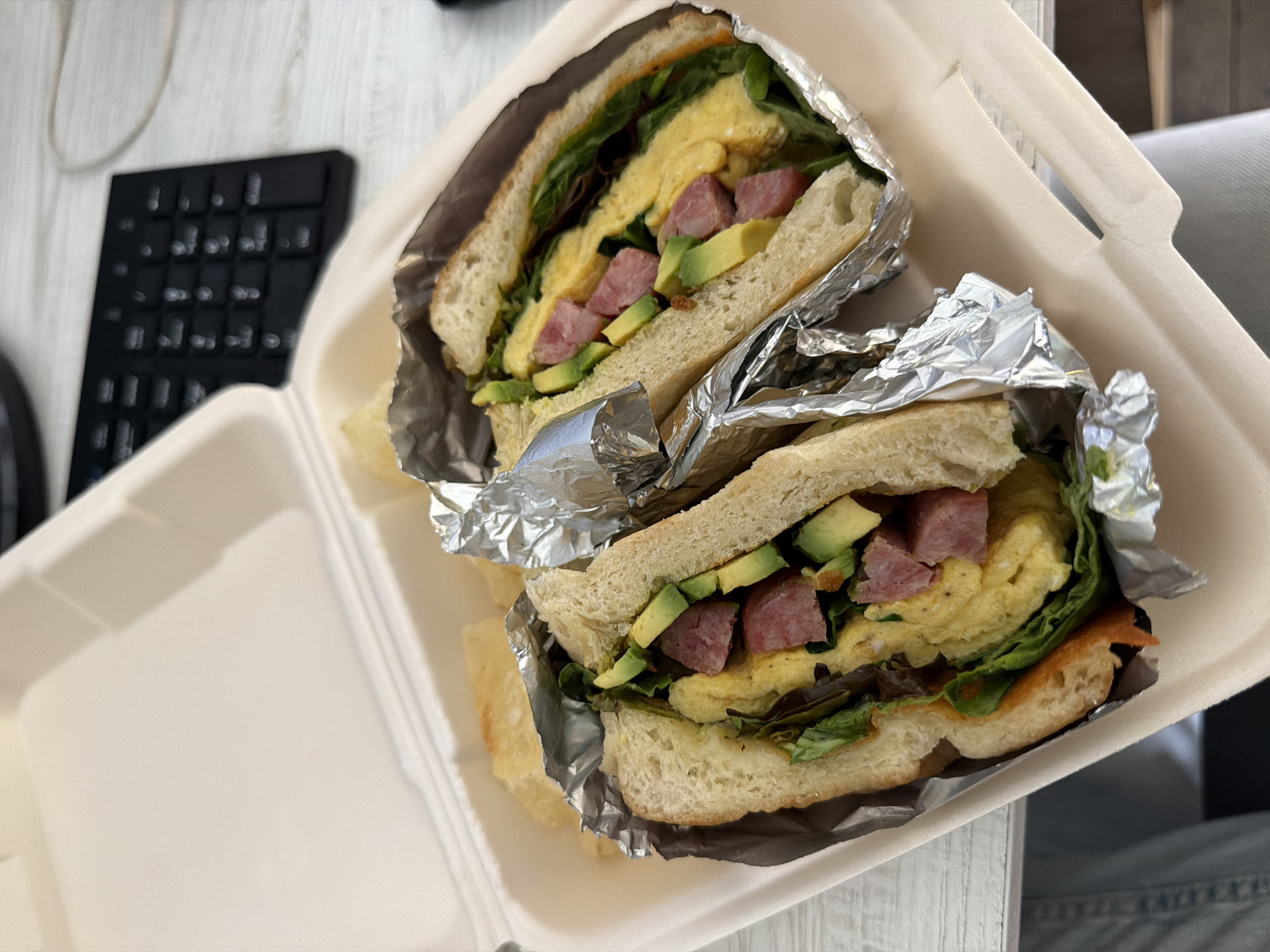 The image shows a takeout container with a sandwich cut in half. The sandwich contains bread, lettuce, avocado, diced meat, and scrambled egg. The sandwich halves are wrapped in aluminum foil inside a white Styrofoam container. Next to the container is a black computer keyboard. The background is nondescript, suggesting an indoor setting. There is no information in the image to suggest a location in Puebla, Mexico.