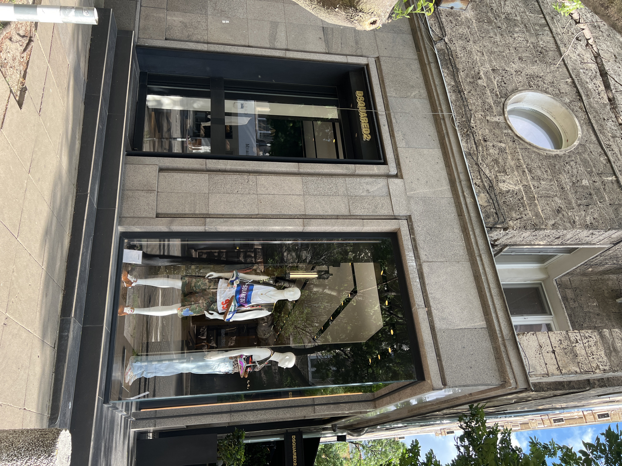 The image shows the storefront of a DSQUARED2 store in Sofia, Bulgaria. The store has two large display windows featuring mannequins dressed in casual clothing. The upper window displays a sign with the brand name "DSQUARED2." The building facade is made of gray stone. To the right of the store is another building with an oval window. The image is taken from a slightly low angle, showing the sidewalk and some trees reflected in the windows.