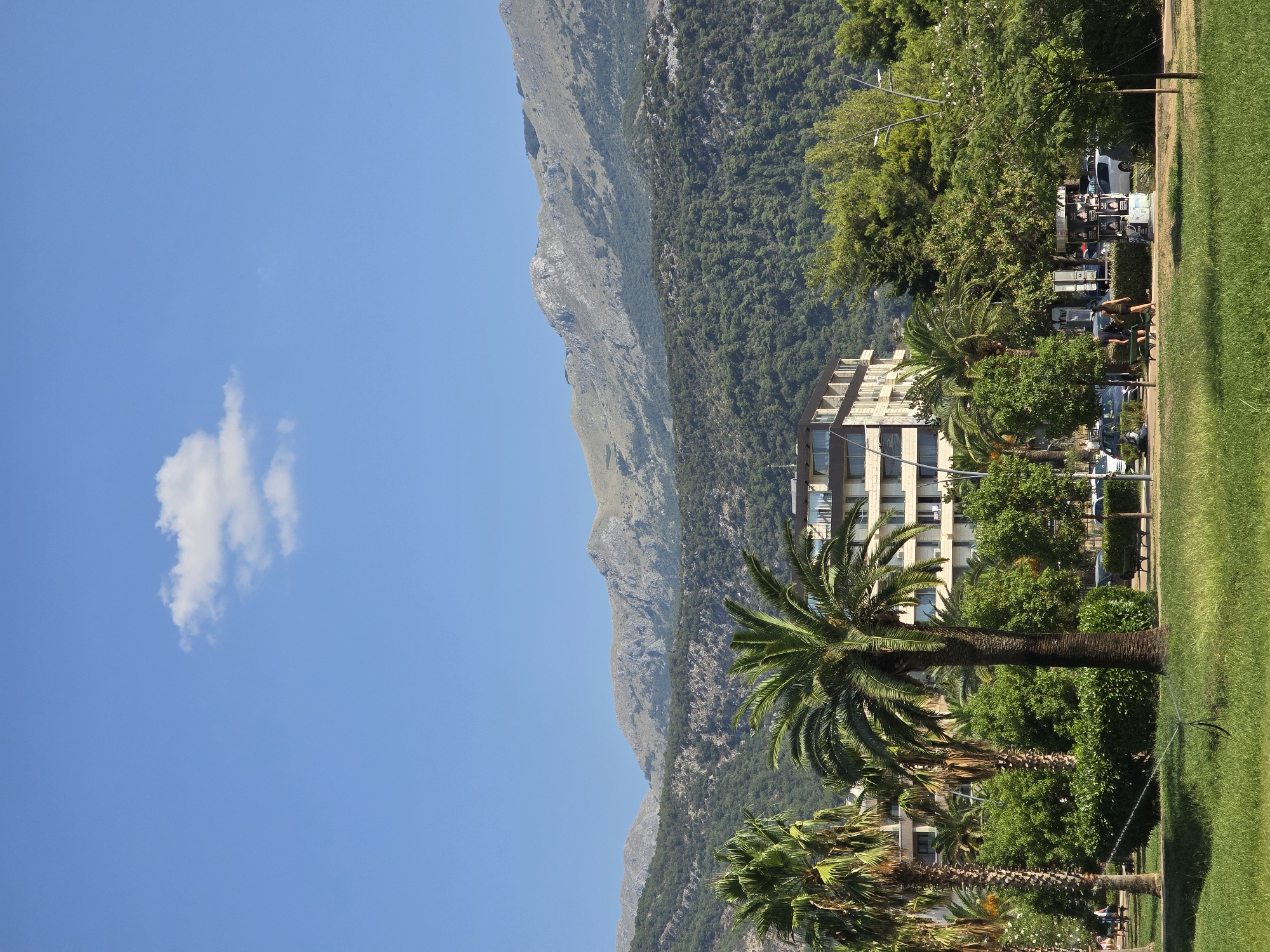 In Budva, Montenegro, on a sunny day, the image captures a vibrant scene with a clear blue sky. A single, fluffy white cloud floats in the upper left quadrant. The middle ground showcases a modern, light-colored, multi-story building with dark windows and balconies, surrounded by lush green trees, including several tall palm trees with prominent fronds. In the foreground, a well-maintained grassy area features a sprinkler system in operation, with a water jet spraying upwards. Beyond the trees, a paved area with benches and parked cars is visible, with a few pedestrians walking. The background is dominated by a majestic mountain range, covered in dense green foliage on its lower slopes and transitioning to rocky, arid terrain towards the peaks. The overall impression is one of a beautiful, sunny day in a resort town, with a blend of urban development and natural beauty.