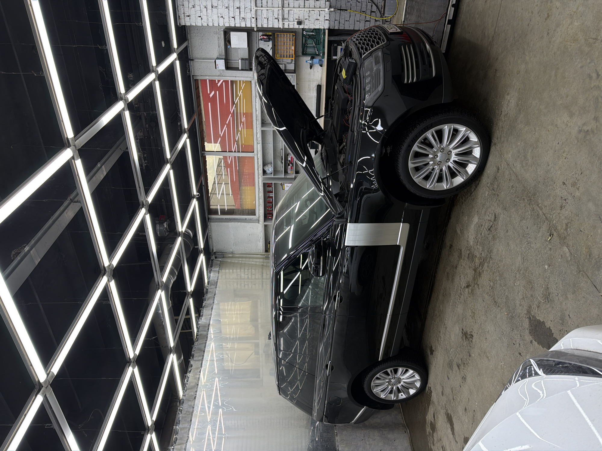 The image shows a black Range Rover with its hood open, parked inside a car detailing shop. The shop has a grid of bright fluorescent lights on the ceiling and white brick walls. A window reveals a red and yellow design on an adjacent wall. Various tools and equipment are visible near the car's open hood. Part of a white car is seen in the lower right corner. The location appears to be Krasnoyarsk, Russia.