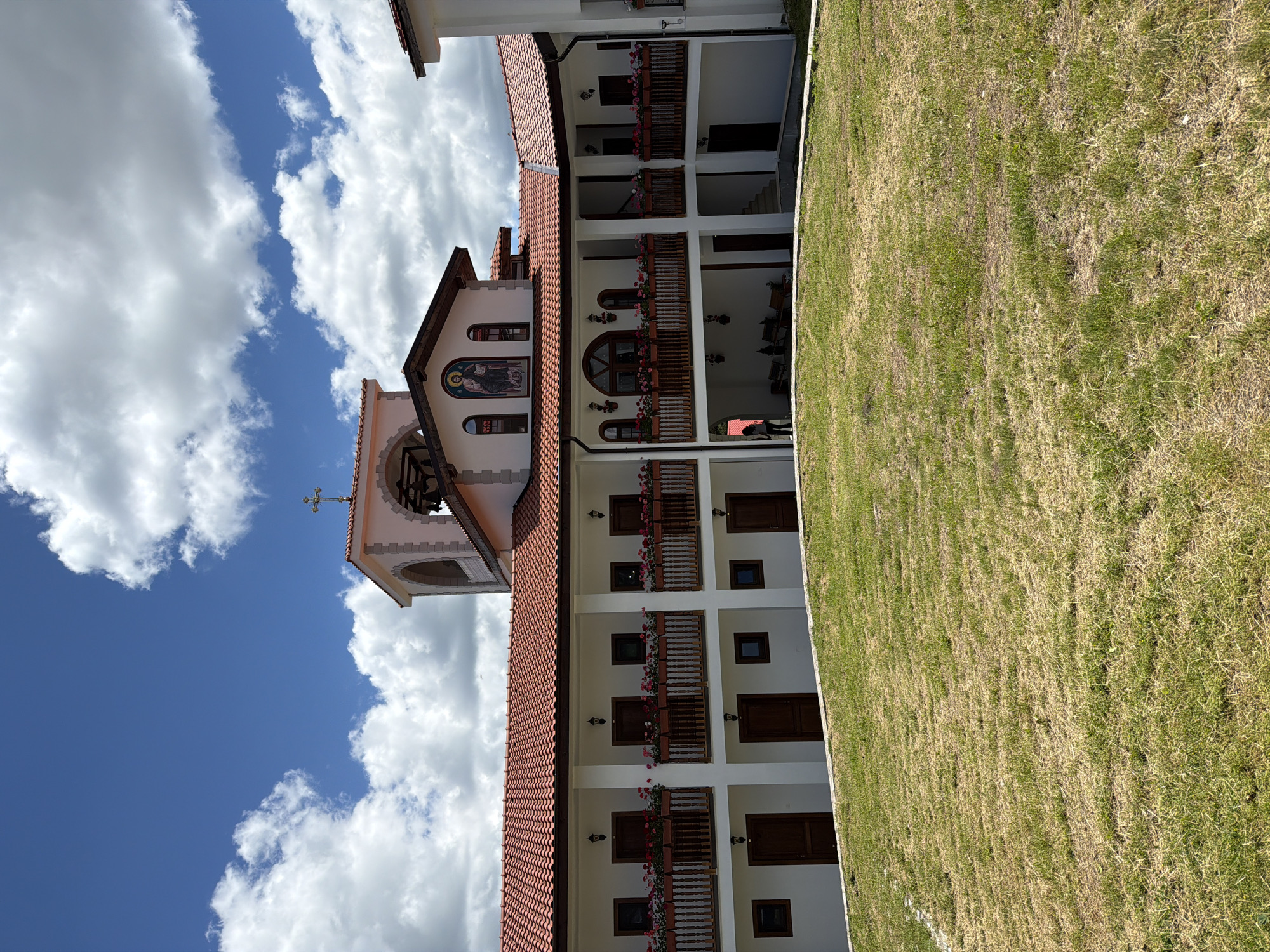 An exterior, eye-level, full shot captures a multi-story, white-walled building with a red-tiled roof under a partly cloudy blue sky. The building features numerous windows and doors, with balconies adorned with flower boxes. A bell tower with a cross on top is visible at the center of the structure. The foreground is occupied by a sloping, dry, grassy hill. A single person, a woman, is visible standing at the bottom level of the building.