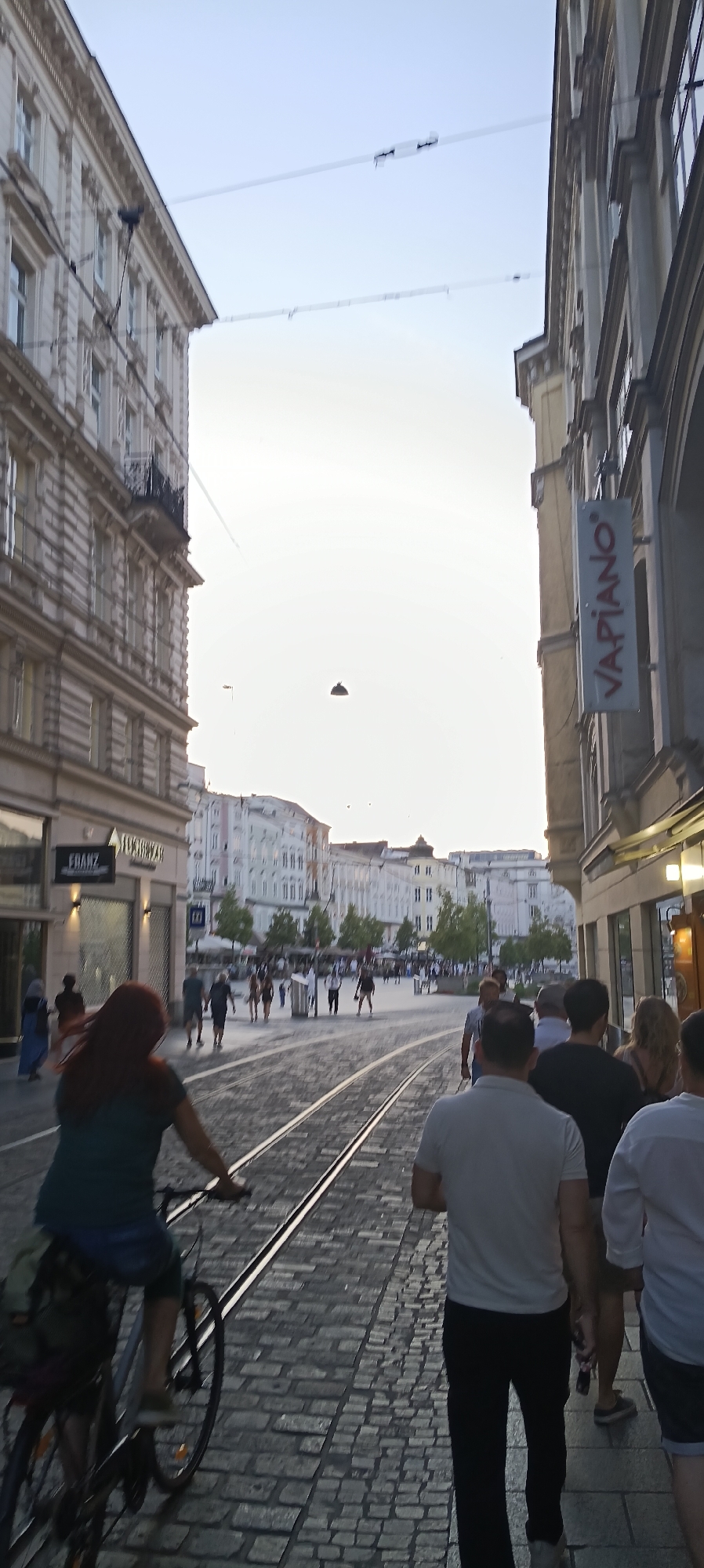 The image depicts a street scene in Linz, Austria, likely on a warm day, judging by the attire of the people present. The perspective is from ground level, looking down a cobblestone street lined with buildings. In the foreground, on the left, a person is riding a bicycle with red hair and a dark green top. The bicycle has a basket on the back. To the right of the cyclist, several individuals are walking away from the camera.
The street features tram tracks, indicating it is a route for public transportation. The buildings on both sides of the street are several stories tall, with a mix of architectural styles. On the left, the building appears to be older, with ornate detailing and balconies. On the right, a building has a sign that reads "VAPIANO" vertically. Further down the street, more buildings are visible, along with trees and outdoor seating areas, suggesting commercial establishments like restaurants or cafes. The sky is clear and light, indicating daytime, possibly late afternoon or early evening. Several power lines or tram wires are visible spanning the street. The overall impression is of a busy urban environment with people engaged in leisurely activities.