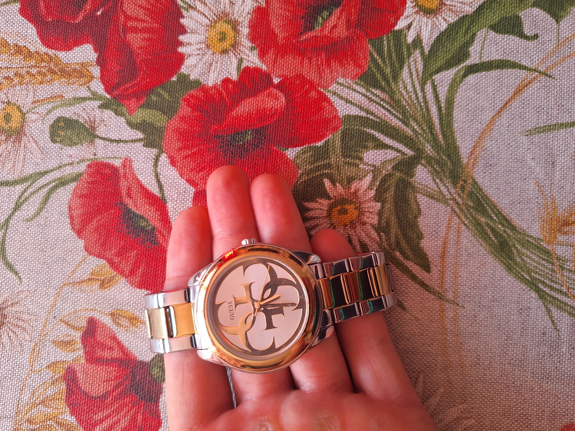 A close-up overhead shot captures a person's hand holding a gold and silver Guess watch against a floral patterned fabric. The watch face features a large, stylized "G" in gold, with the word "GUESS" printed above it. The fabric background is a light beige with a repeating pattern of red poppies, white daisies, and green foliage. The lighting is natural and casts subtle shadows. There is no information available about the location in Varna, Bulgaria, or any specific actions or interactions occurring in the scene.